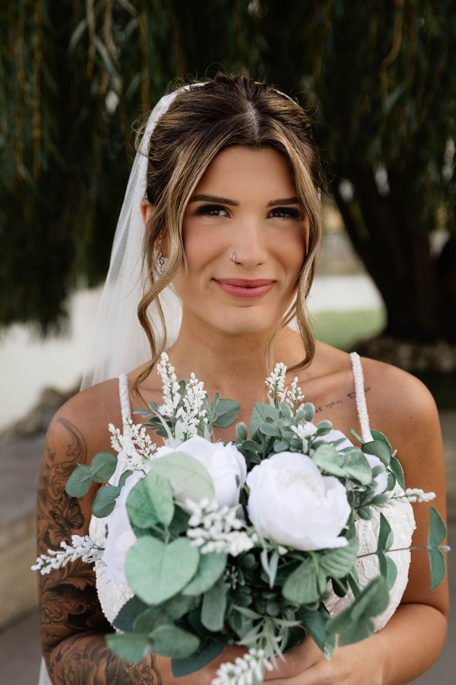 Beautiful bride with tattoos holds a white peony and eucalyptus bouquet wearing a veil outdoors.
