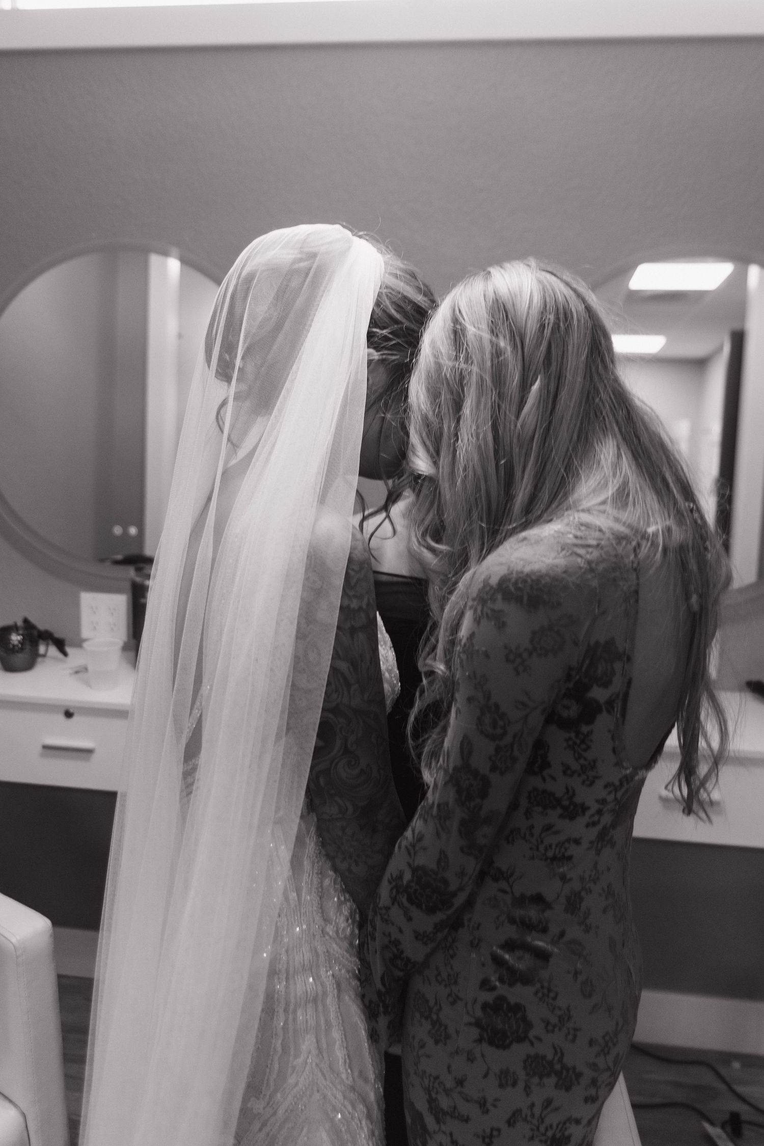 Black and white photo of bride in veil being helped by woman in floral dress in bridal suite.