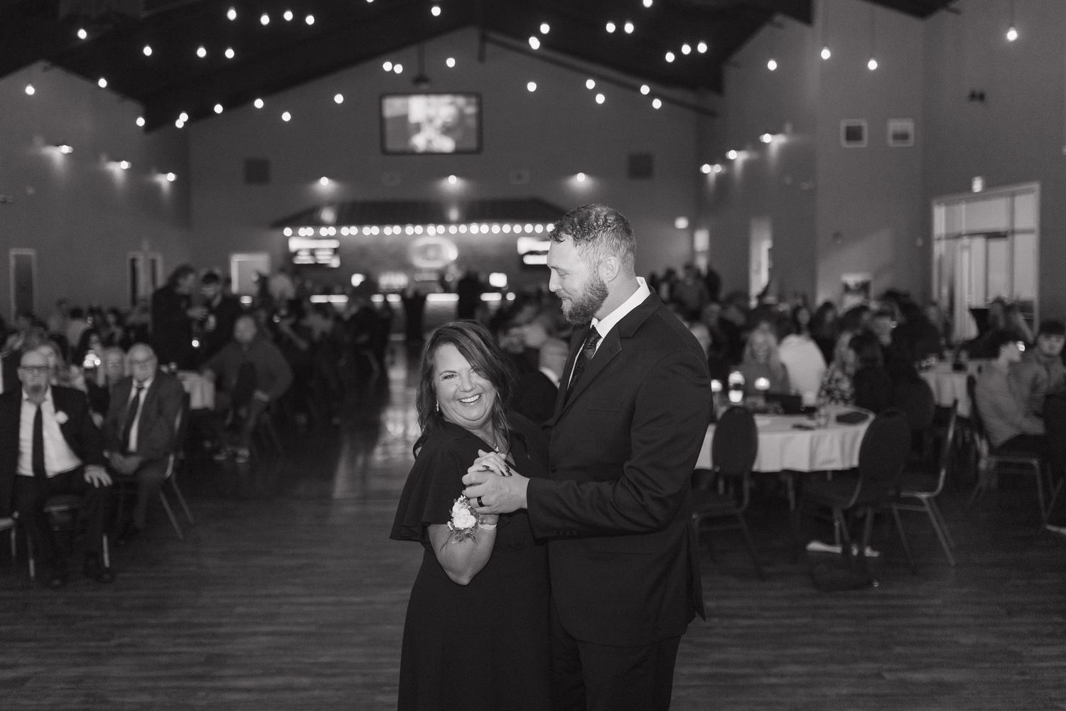Black and white photo of a couple sharing a dance at an elegant wedding reception venue with string lights.