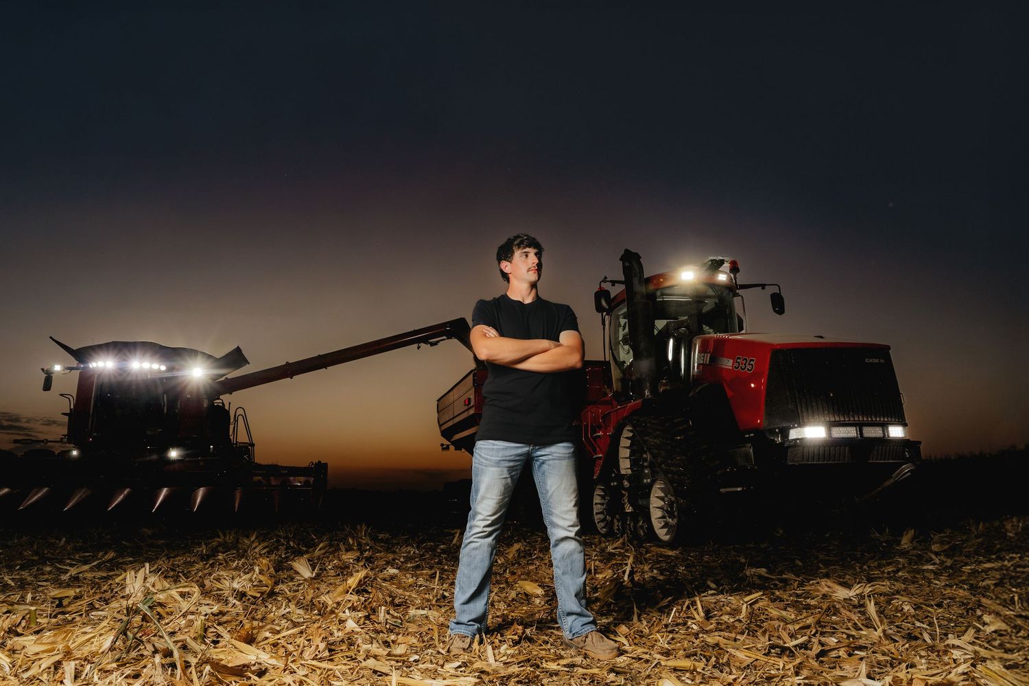 Man stands with arms crossed beside a Case IH tractor and grain cart at night in field.
