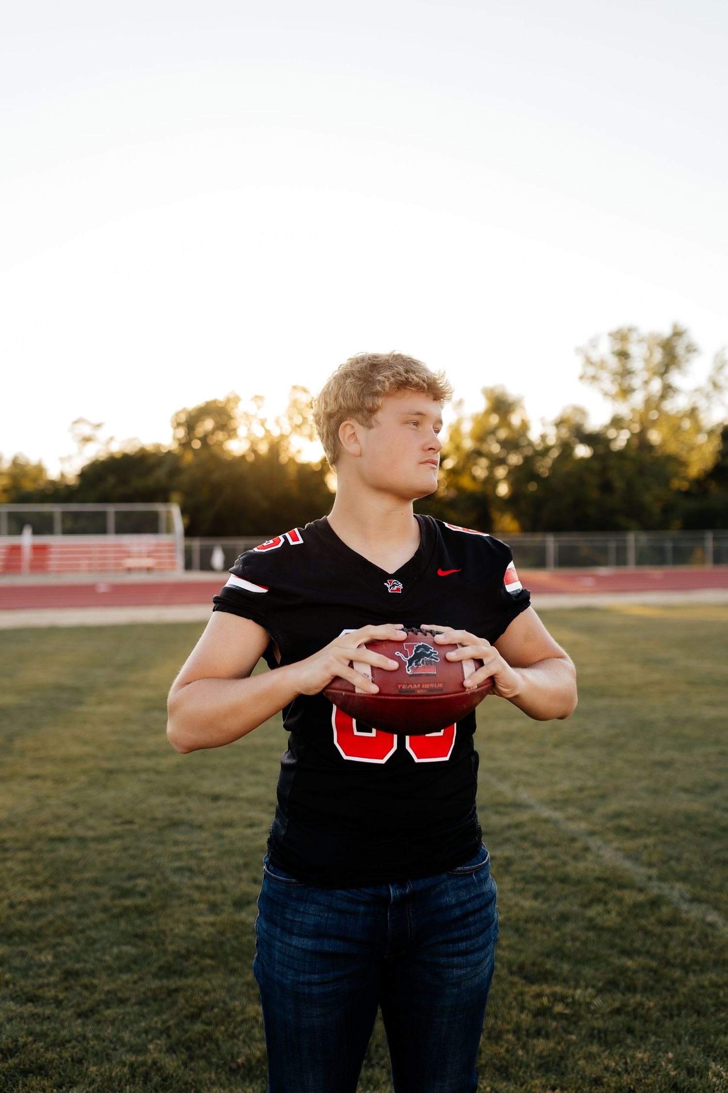 Young football player in black jersey holding a football on a field at golden hour.