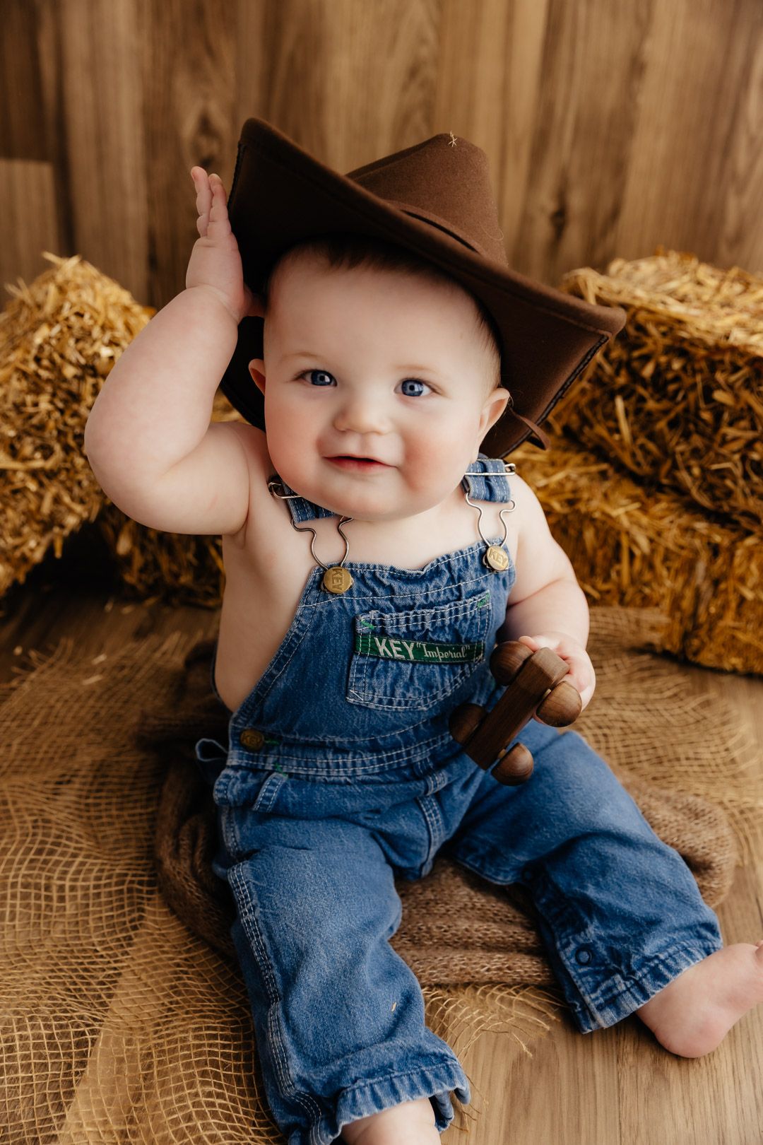 Happy baby in denim overalls and brown cowboy hat sitting on burlap near hay bales in rustic setting.