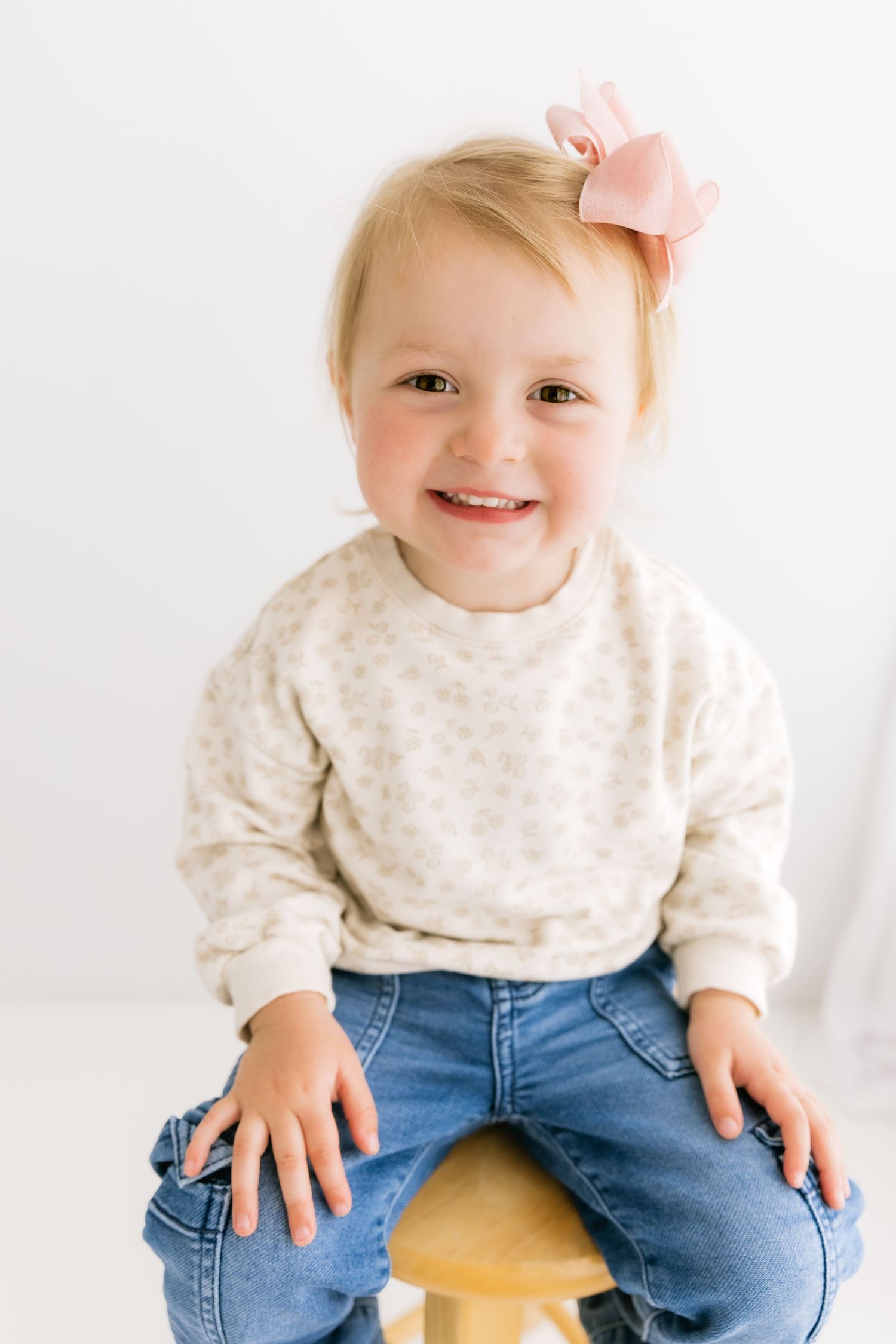 Sweet toddler girl with pink hair bow sits on wooden stool smiling in white photography studio.