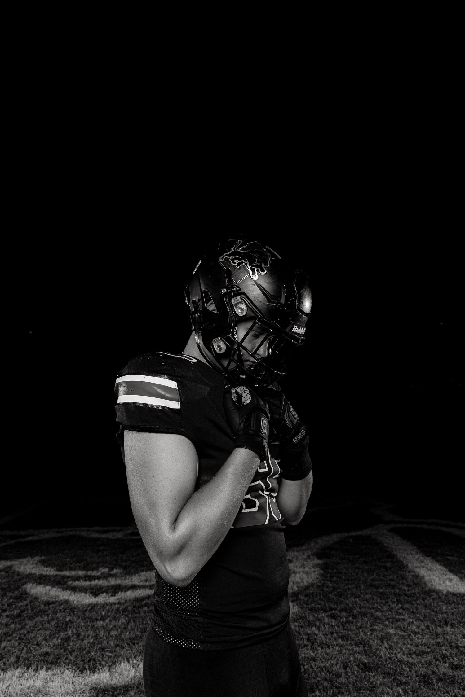 Black and white portrait of a football player in full gear and helmet standing on dark field.