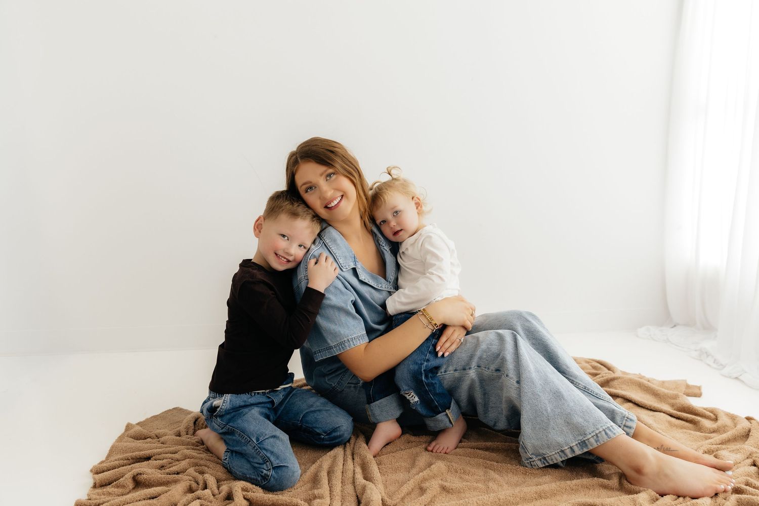 Mother sitting on tan blanket smiling with young son and daughter leaning against her sides.