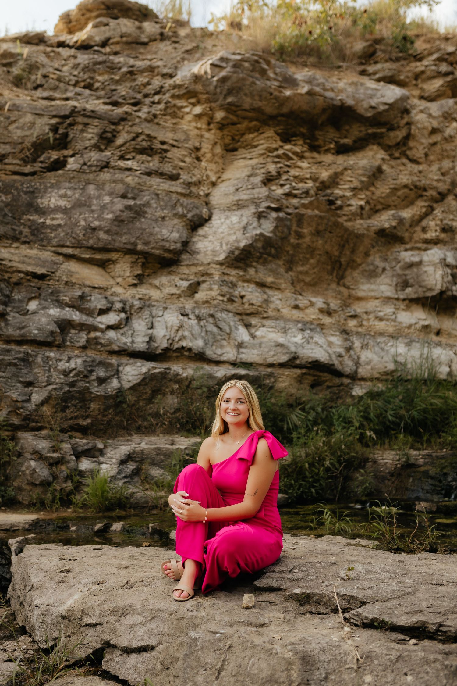 Smiling blonde woman in a hot pink jumpsuit sitting on rocks in front of a natural stone cliff.