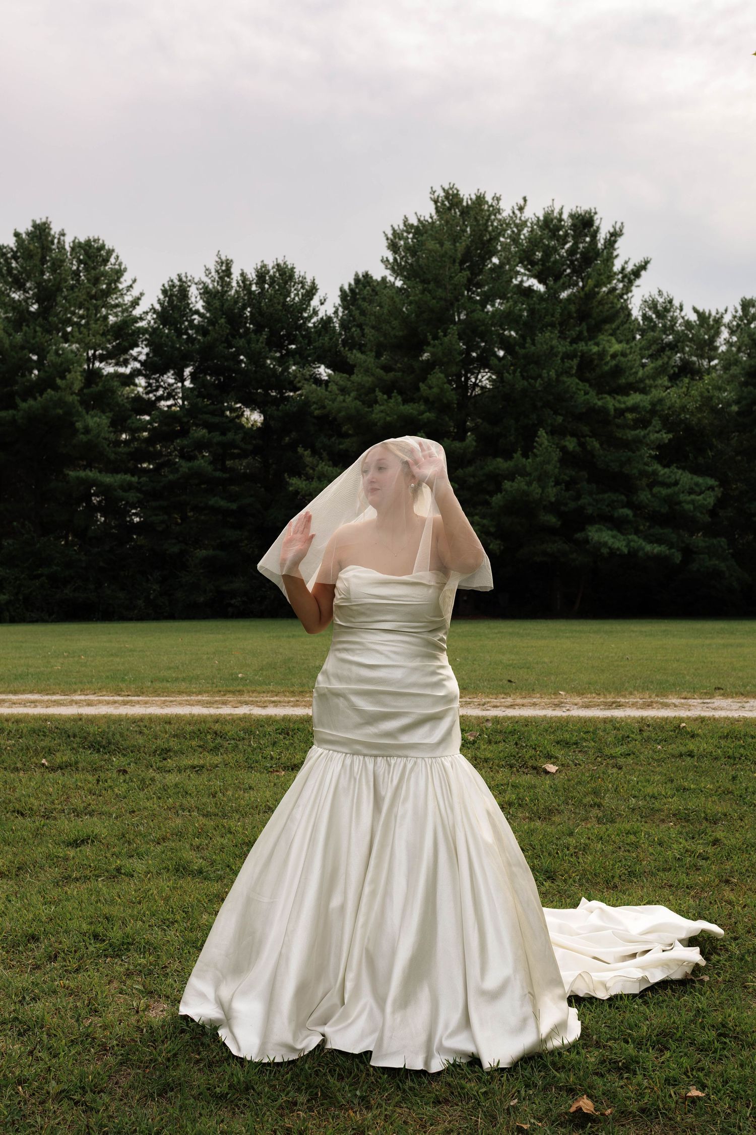 Elegant bride in white mermaid gown adjusts her veil while standing on a green lawn before trees.
