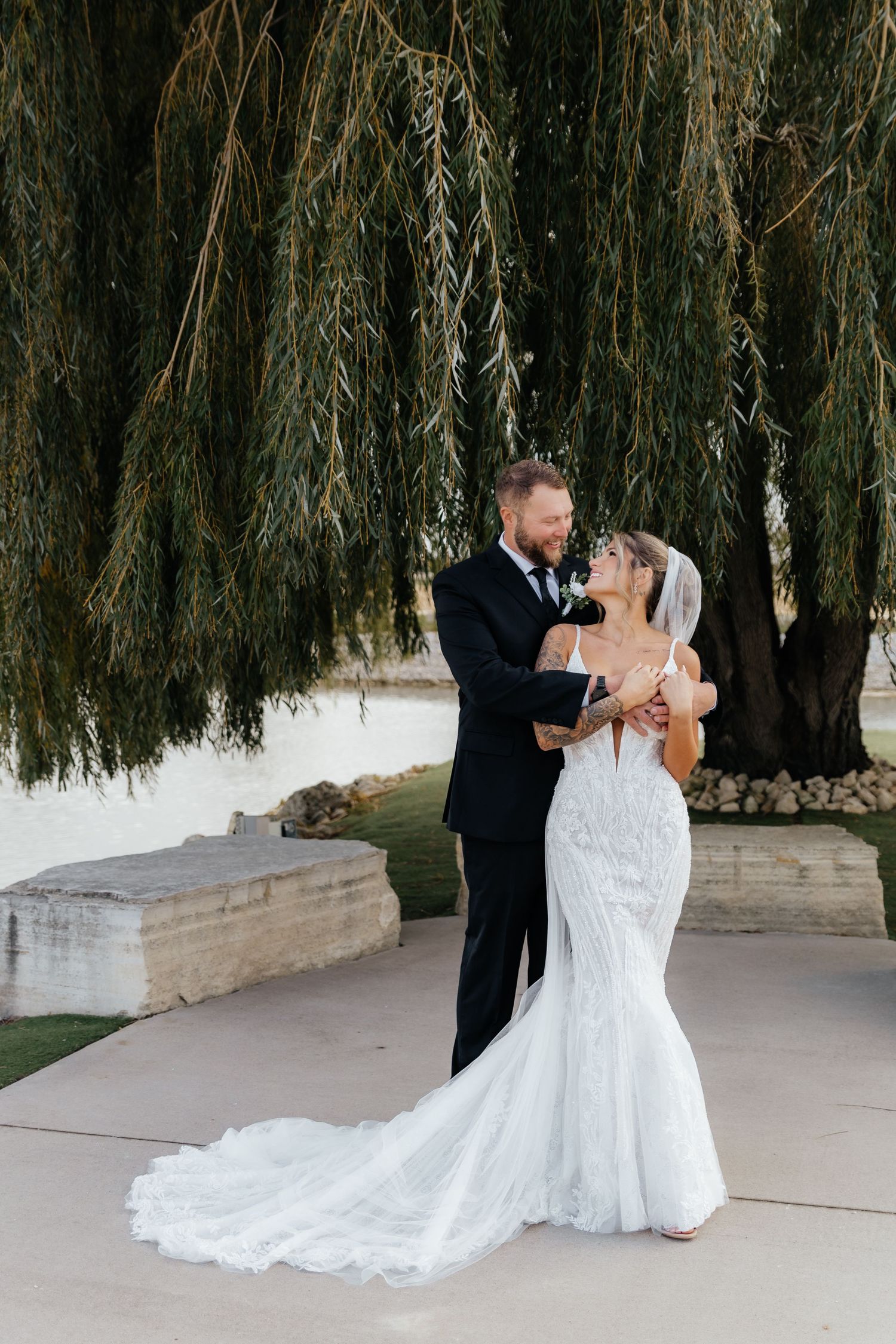 Bride in fitted lace gown with long train stands with groom under weeping willow at outdoor venue.