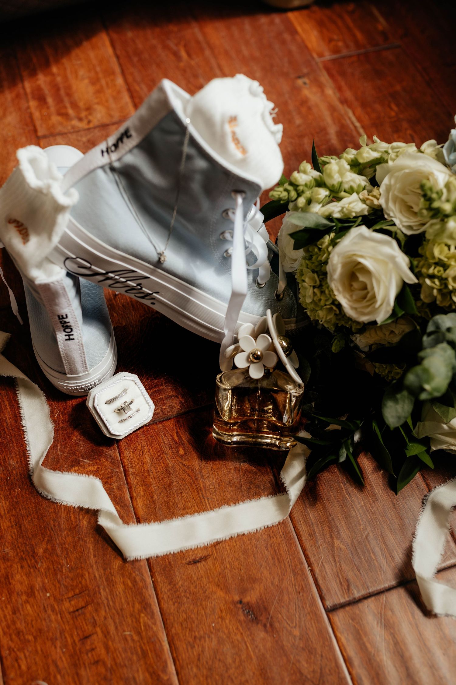 White Converse sneakers labeled Hope beside bridal bouquet, perfume bottle, and ring box on wood floor.