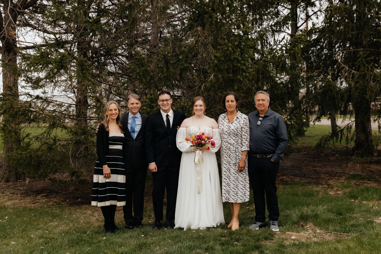 Six family members pose with bride and groom outdoors near tall evergreen trees.