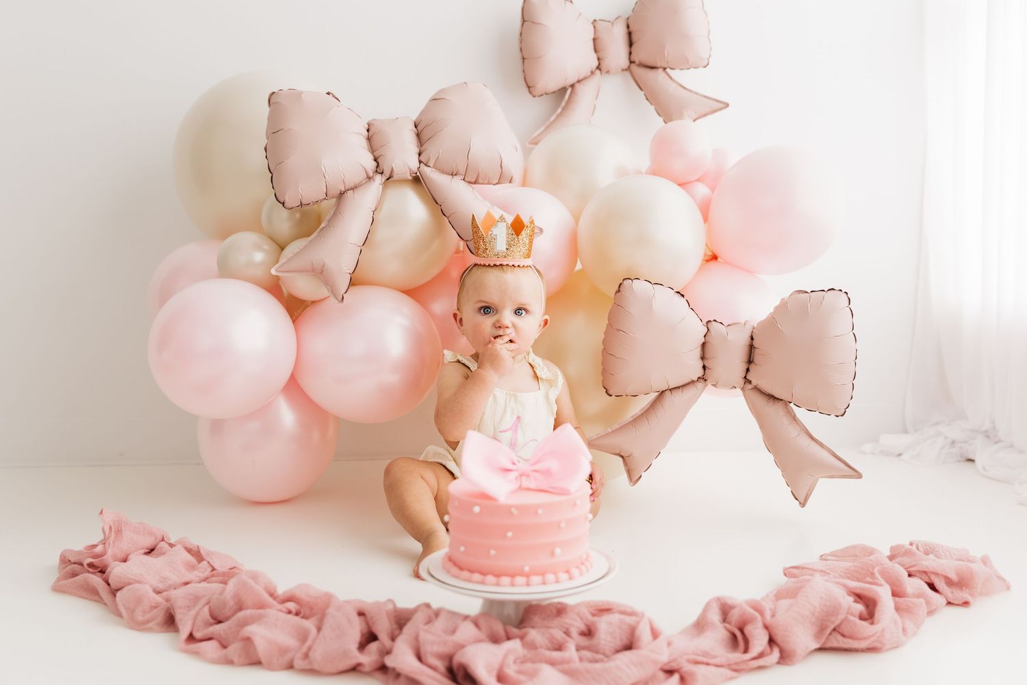 Baby girl in crown celebrates first birthday with pink cake, bow balloons, and pink ribbon decor.