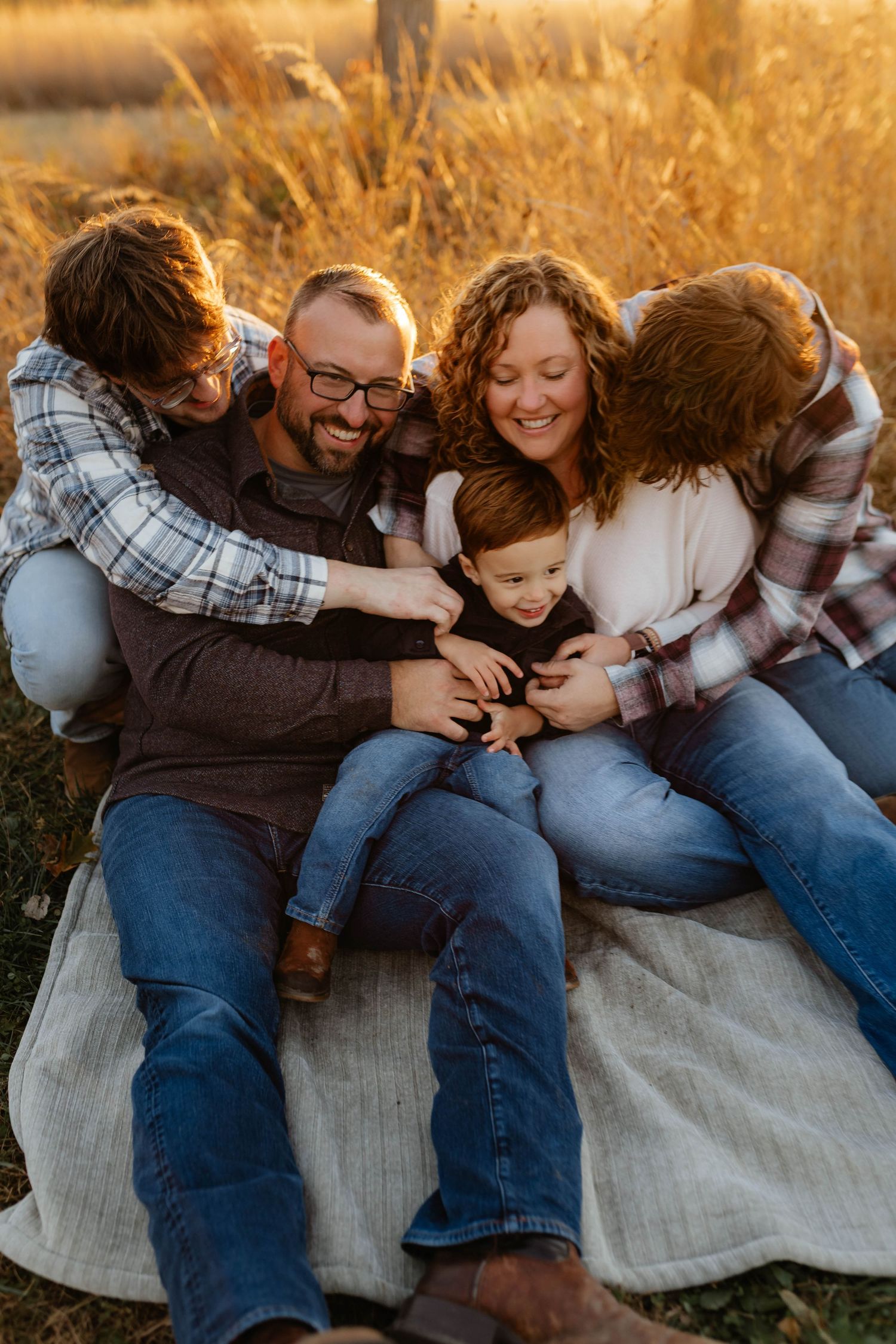Family of five sitting on blanket in golden field laughing together at sunset.