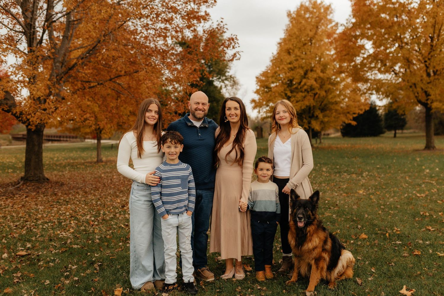 Family of six with German Shepherd dog posing together in autumn park with vibrant orange and yellow foliage.