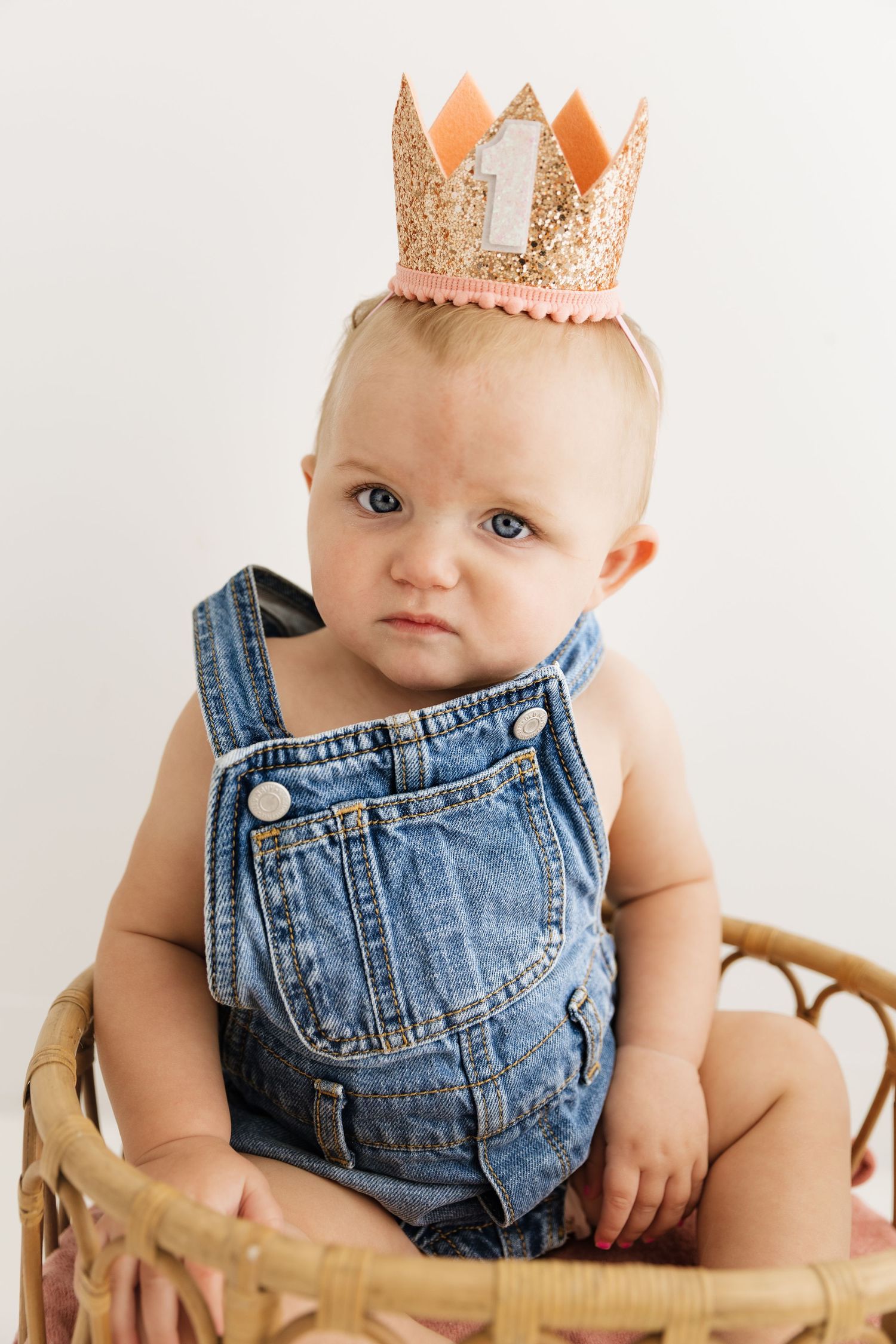 Adorable baby in denim overalls wearing glittery number one birthday crown sits in wicker basket.