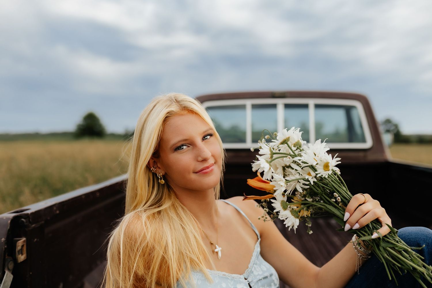 A blonde girl sits in a truck bed holding white daisies, smiling softly against a cloudy field backdrop.
