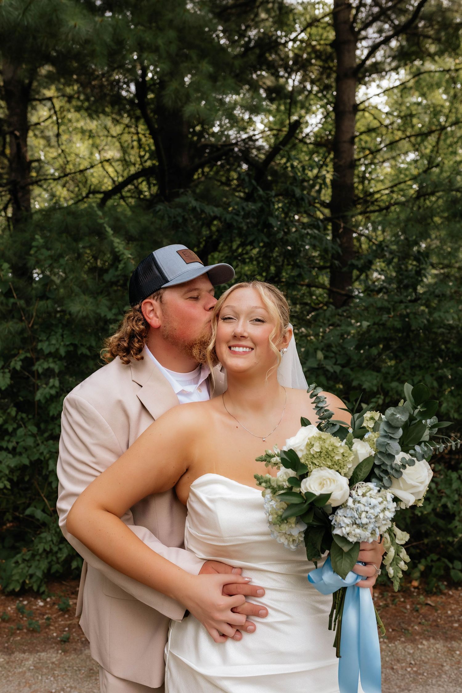 Smiling bride in white strapless gown holds bouquet as groom in tan suit kisses her cheek outdoors.