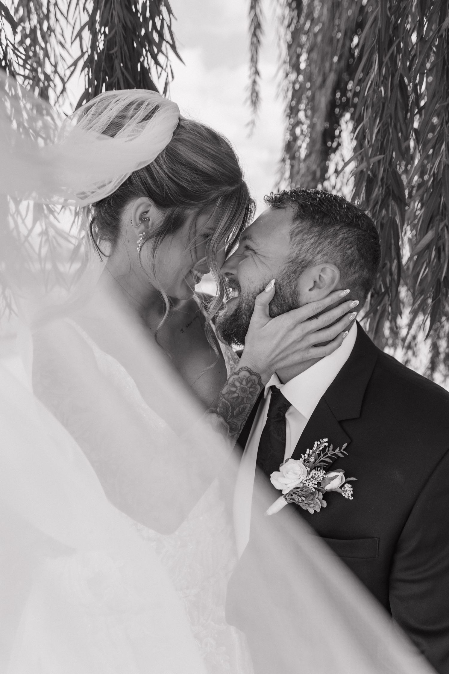 Black and white romantic photo of bride and groom nearly kissing under weeping willow tree branches.