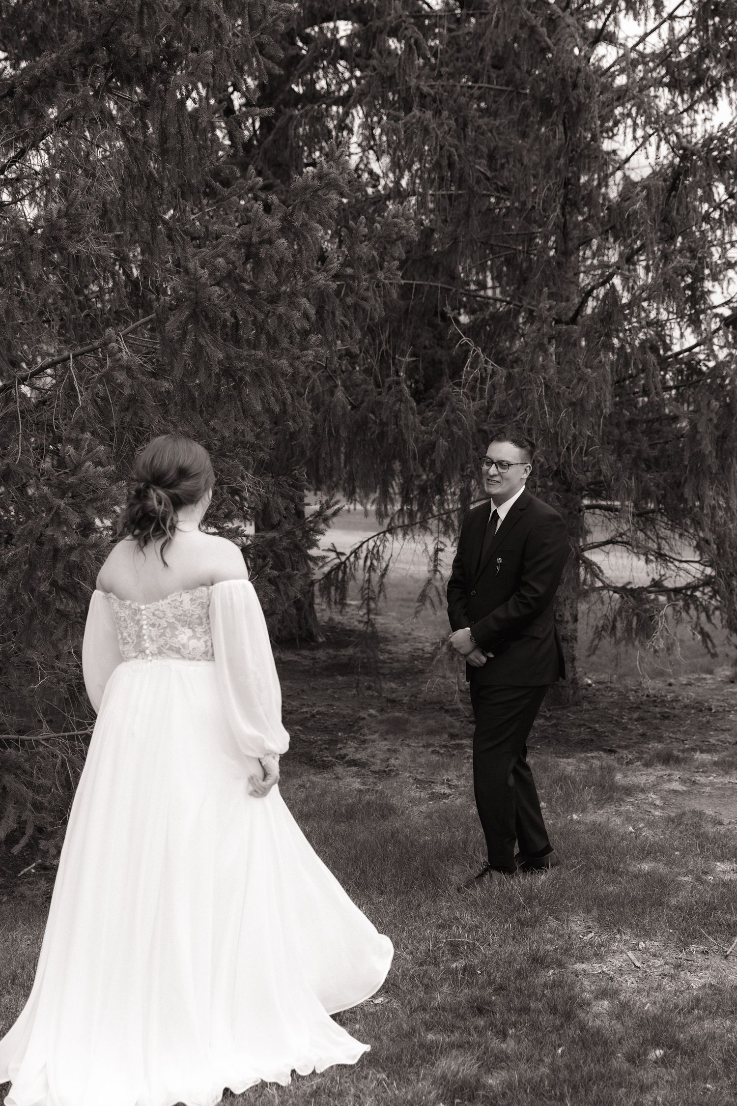 Black and white photo of bride in off-shoulder gown meeting groom for first look under pine trees outdoors.