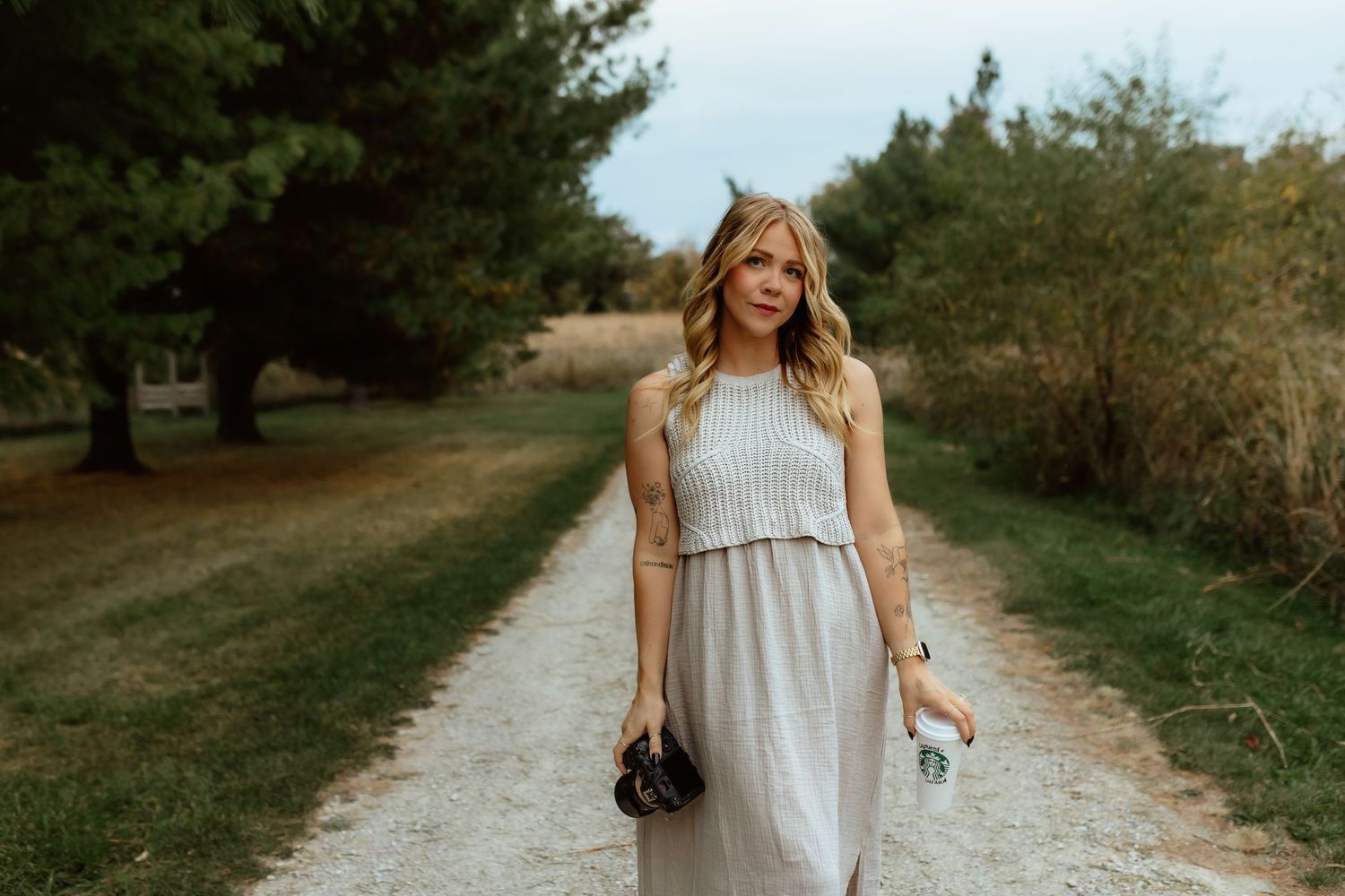 Blonde woman in elegant gray maxi dress walking on a gravel path surrounded by lush green trees.