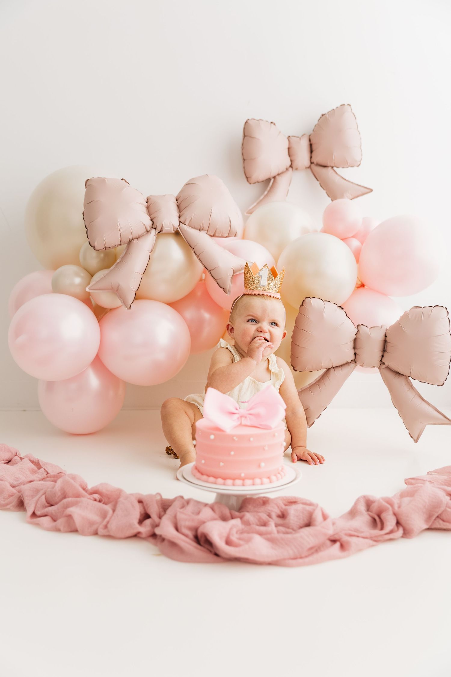 Baby girl in crown smashes pink first birthday cake surrounded by pink and cream bow balloons.