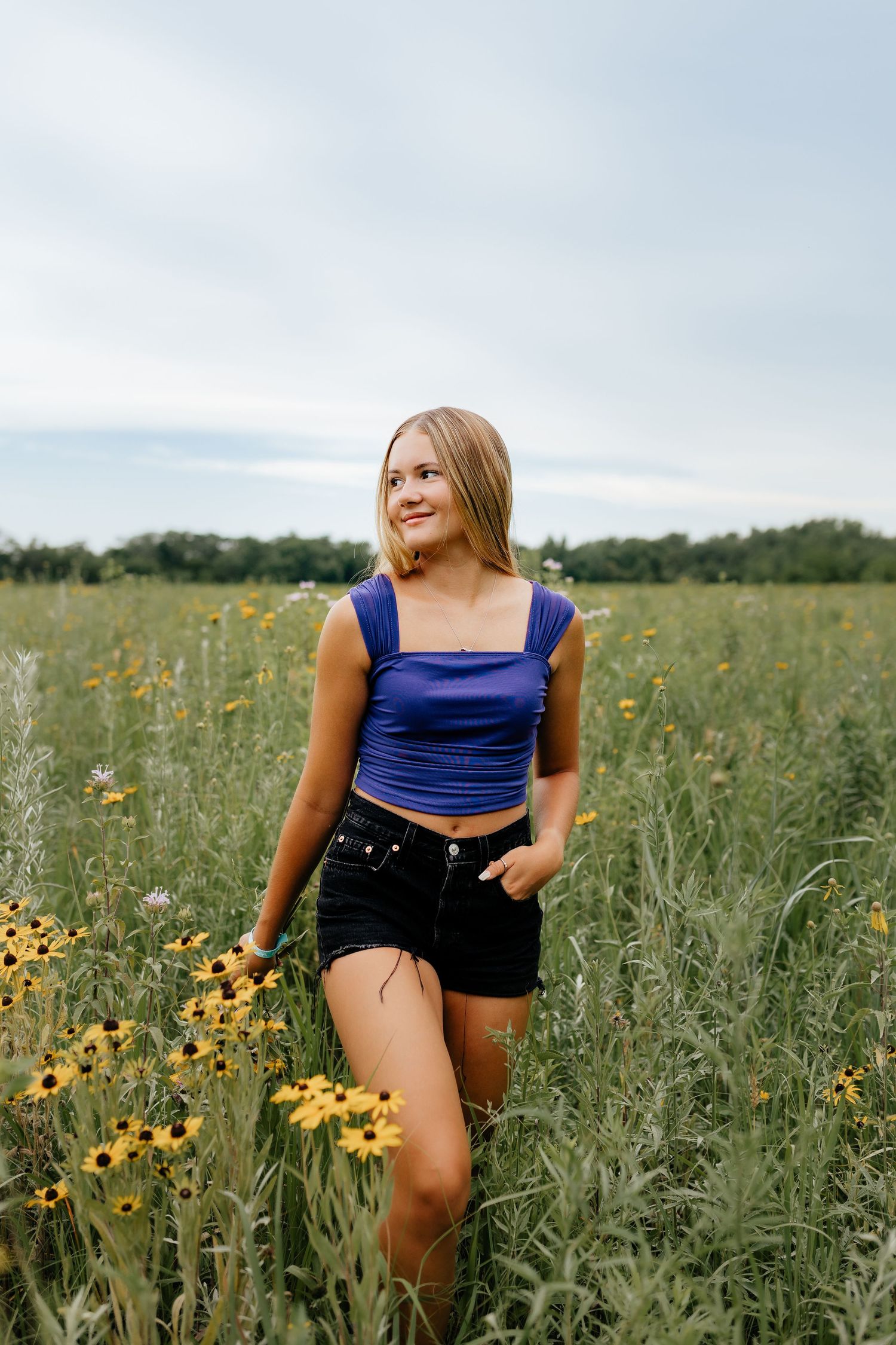 Blonde teen in blue crop top and black shorts stands among yellow wildflowers in green field.