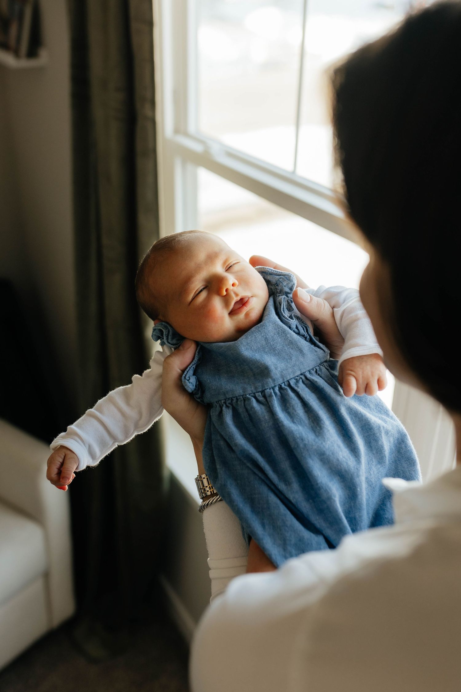 A mother in a white sweater holds her sleeping newborn baby up near a bright window.