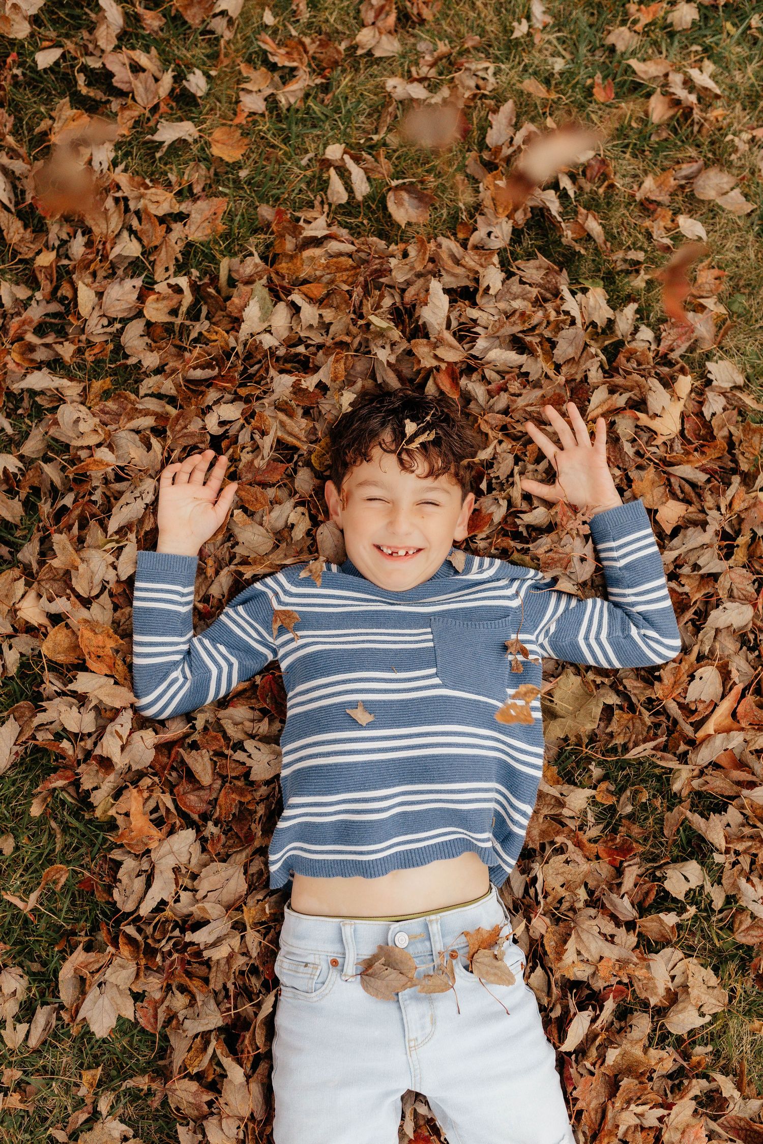 Happy young boy laying in a pile of fallen autumn leaves with arms raised and big smile.