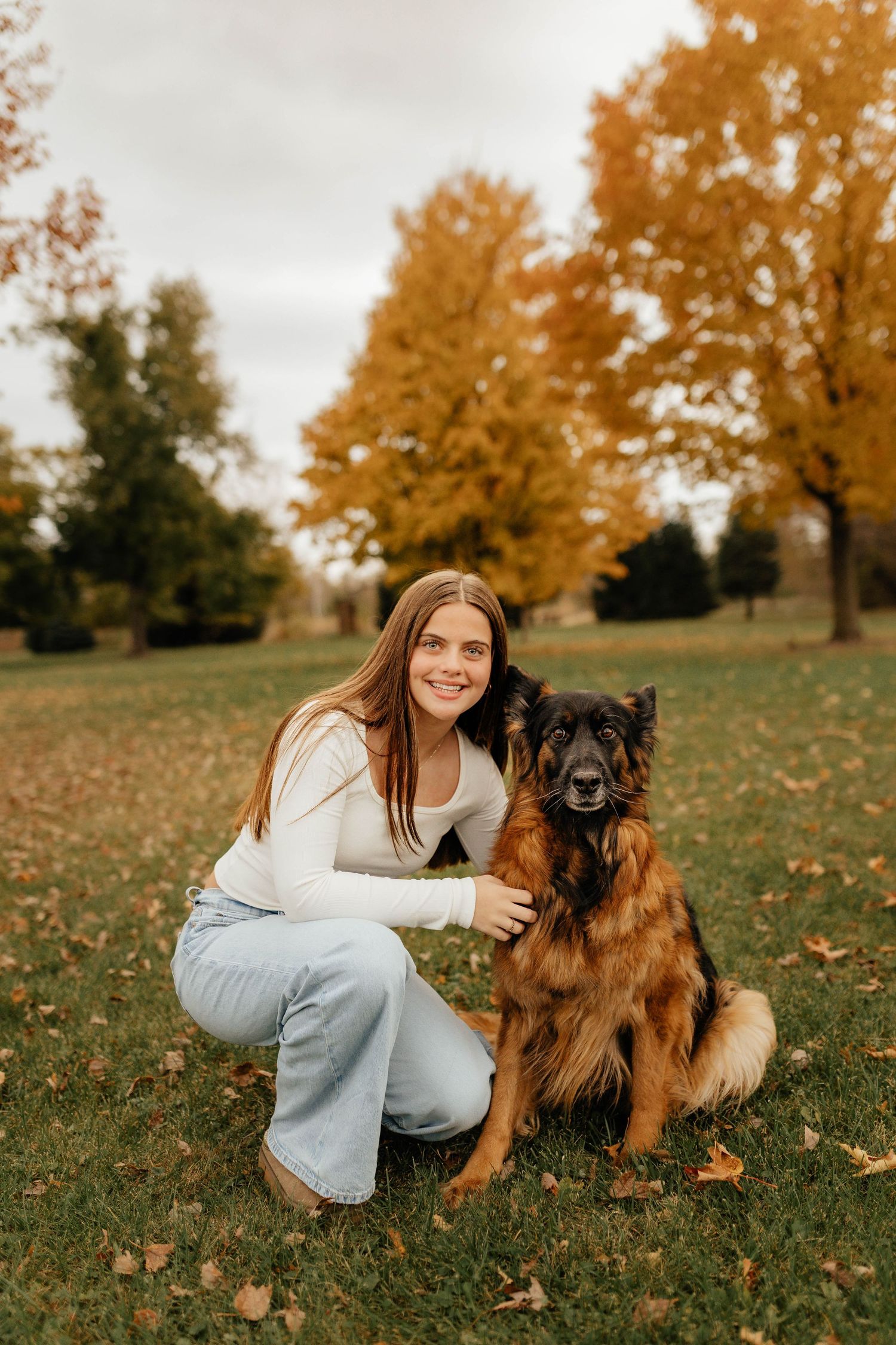 Smiling teenage girl crouching beside a German Shepherd dog on green grass with fall foliage in background.