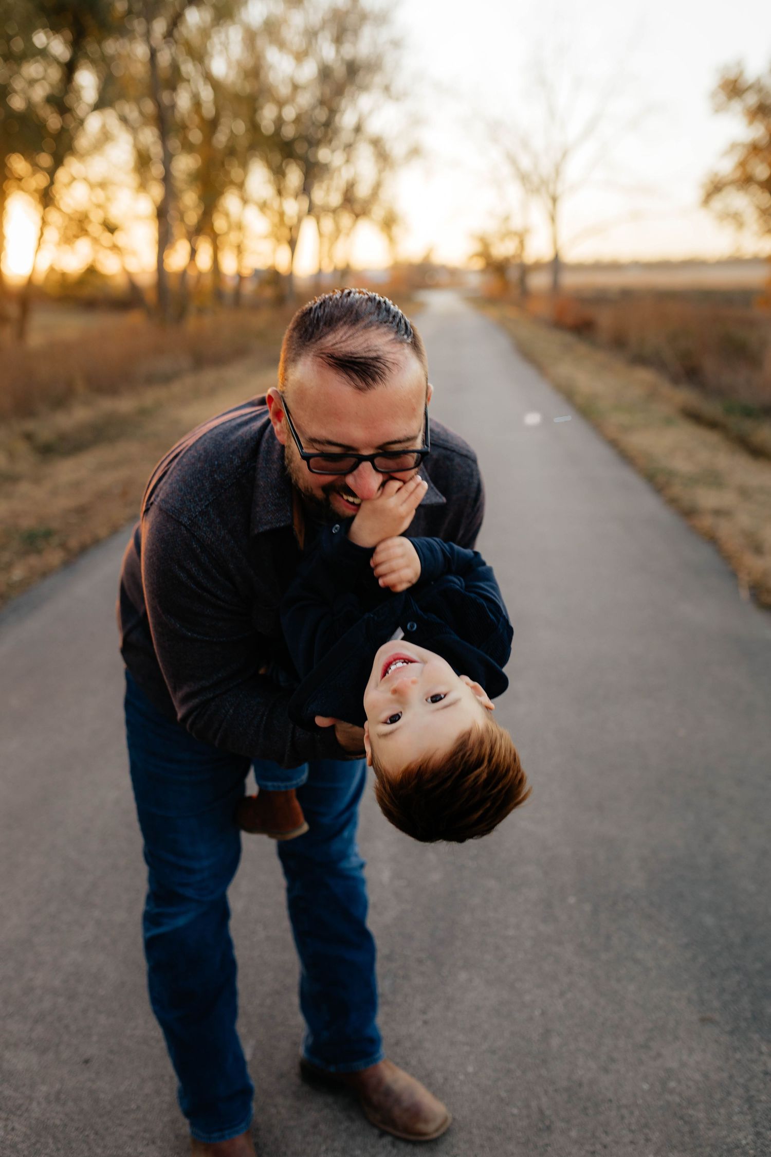 Father playfully holds toddler son upside down on country road at golden sunset.