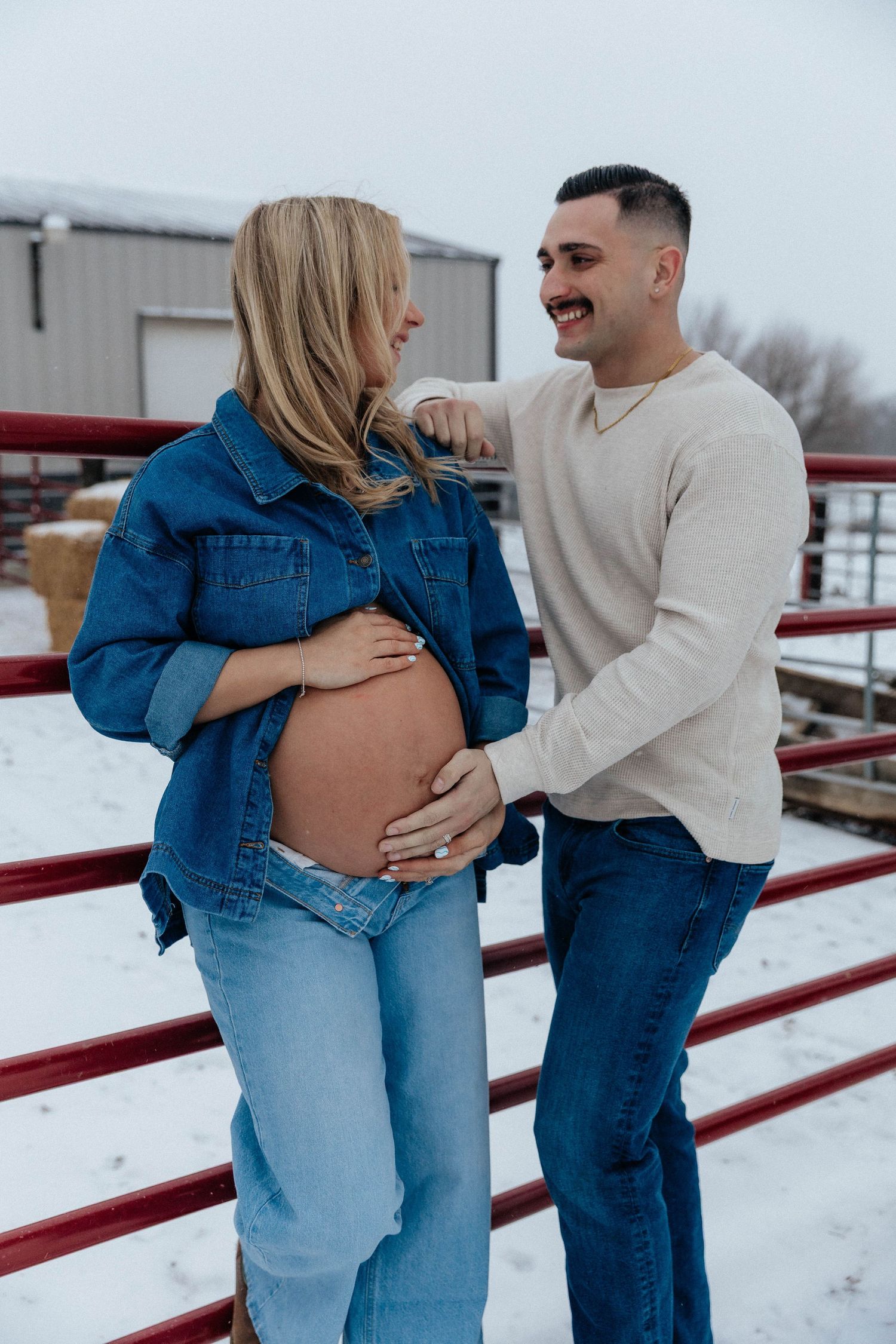 Smiling couple cradles pregnant belly beside red farm fence during winter maternity session.