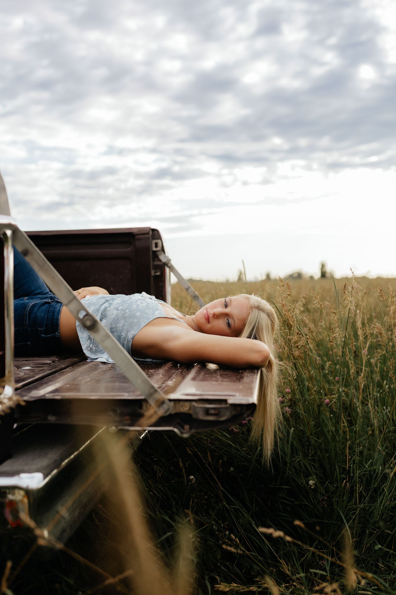 Young blonde woman relaxing in truck bed in grassy field under cloudy sky.
