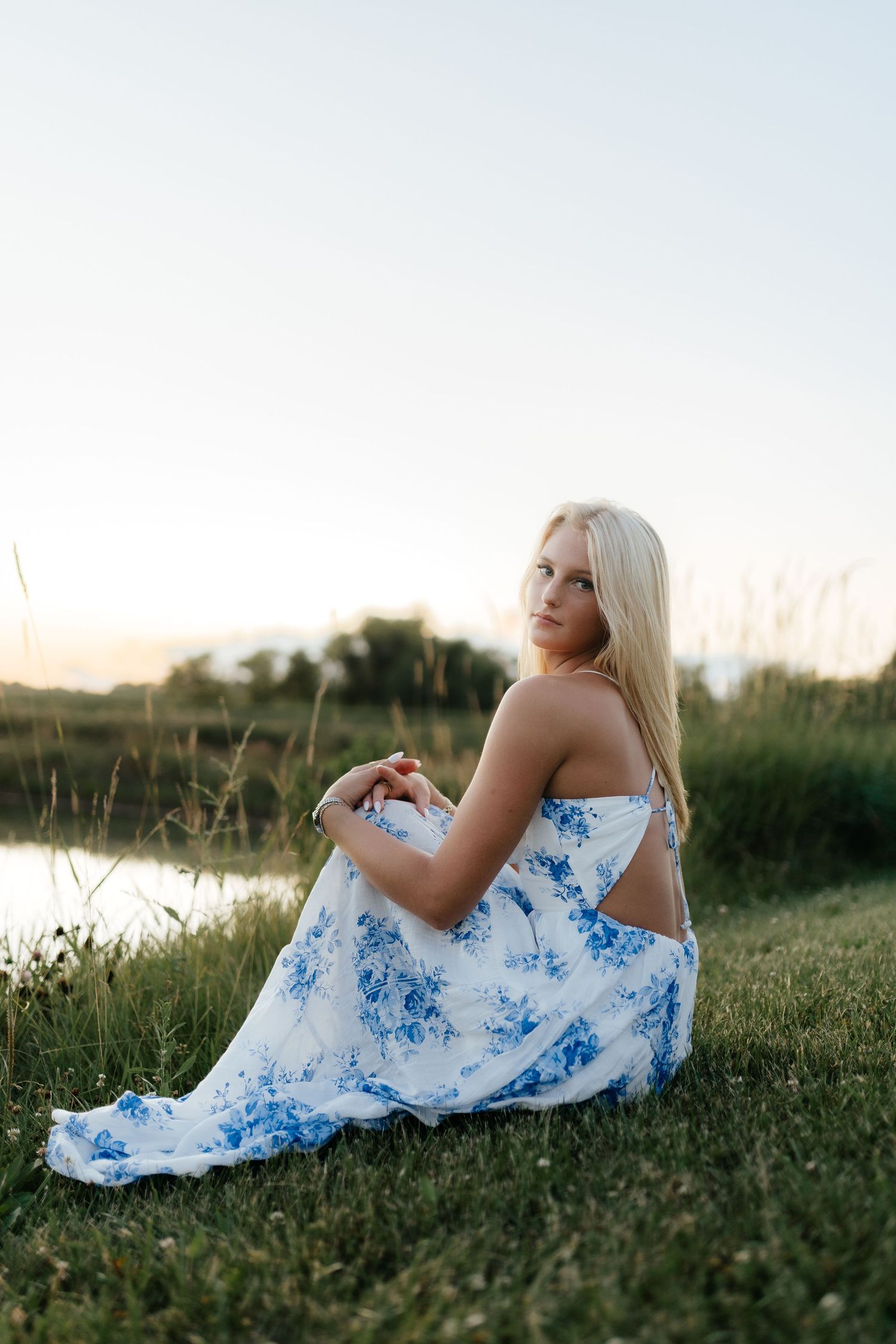 Blonde teenage girl in white blue floral dress sitting on grass by a peaceful pond at sunset.