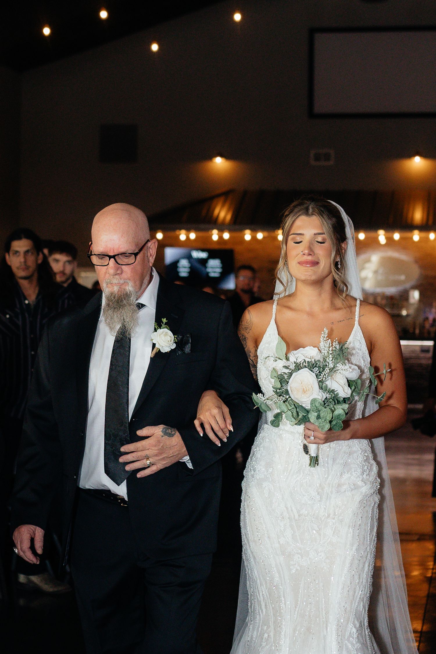 Emotional bride in lace wedding gown walks down aisle with father during wedding ceremony processional.
