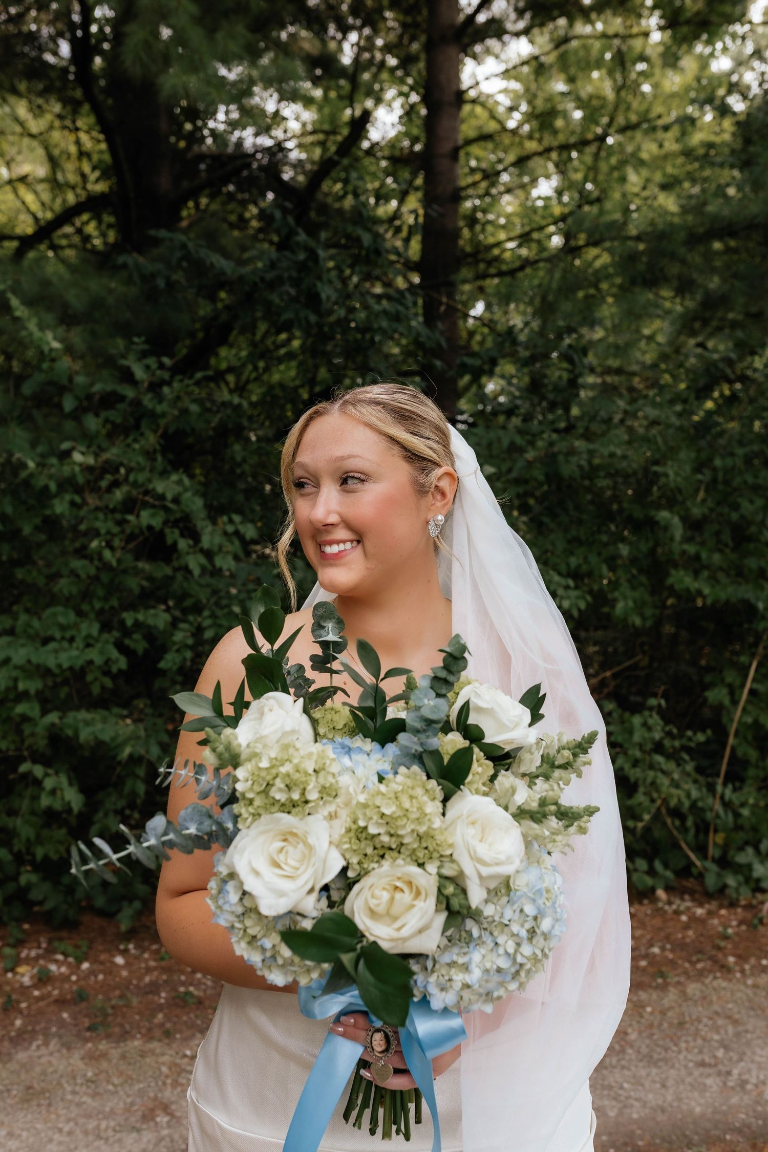 Radiant bride in white strapless gown smiles holding white rose and blue hydrangea bouquet outdoors.