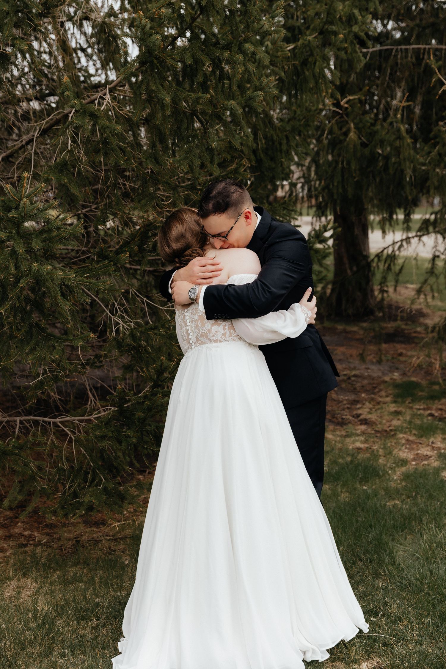 Bride and groom sharing an emotional first look hug beneath pine trees in outdoor wedding portrait.