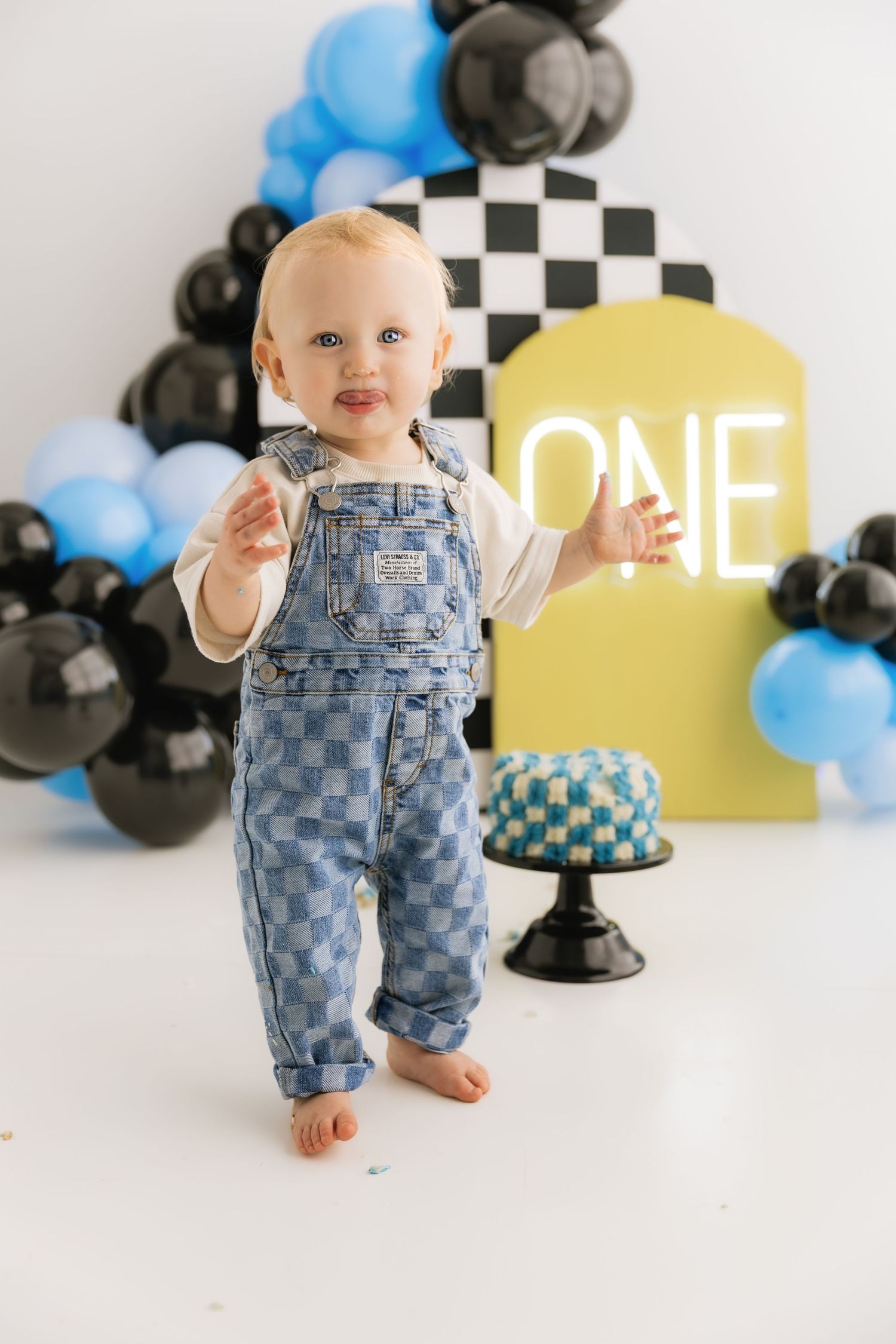 Happy baby boy in checkered denim overalls standing at race car themed first birthday cake smash setup.