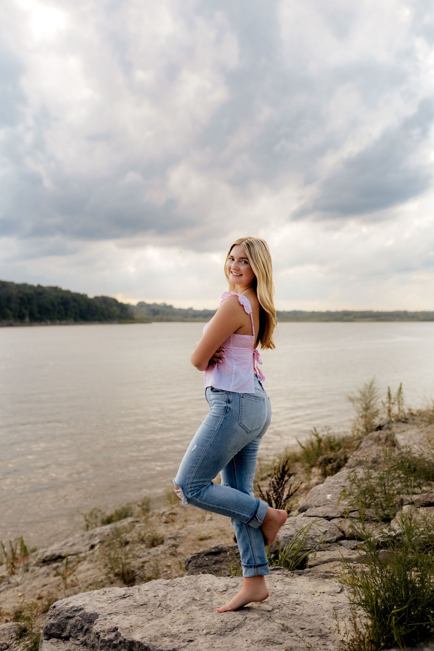 Smiling blonde teen in pink top and jeans posing on rocky lakeshore under cloudy skies.