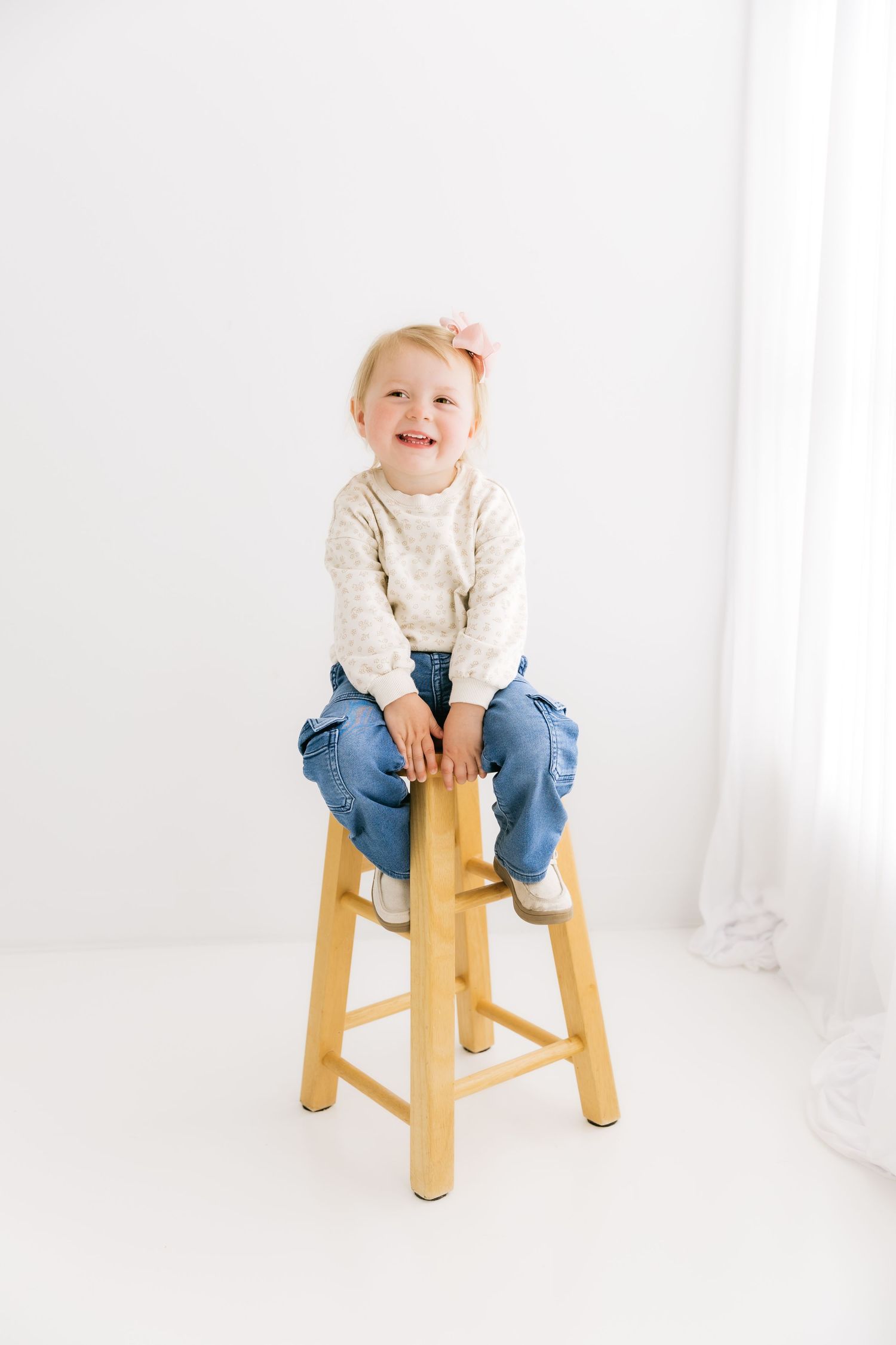 Adorable toddler girl with pink bow sits on wooden stool smiling in white studio setting.