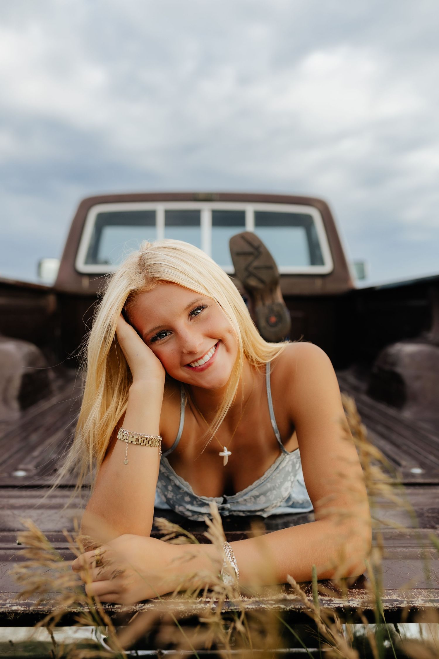 Smiling blonde girl lying in truck bed with cross necklace and bracelet, grass in foreground, cloudy sky.