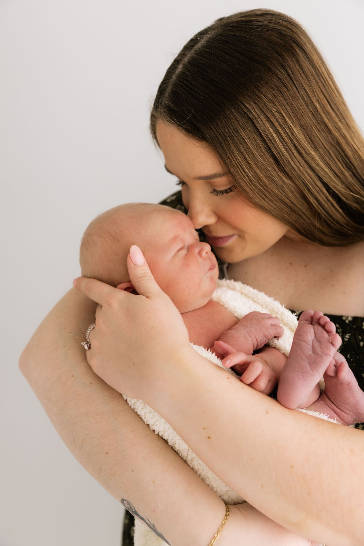 Tender moment of a young mother nuzzling her sleeping newborn baby against a white background.