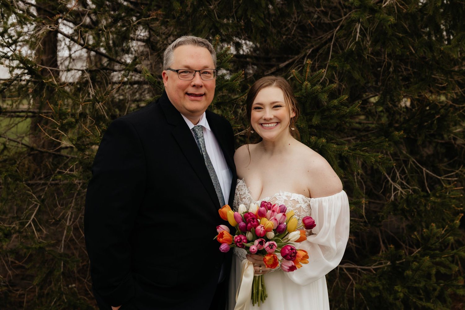 Older man and smiling bride pose together outdoors holding colorful tulip bouquet.