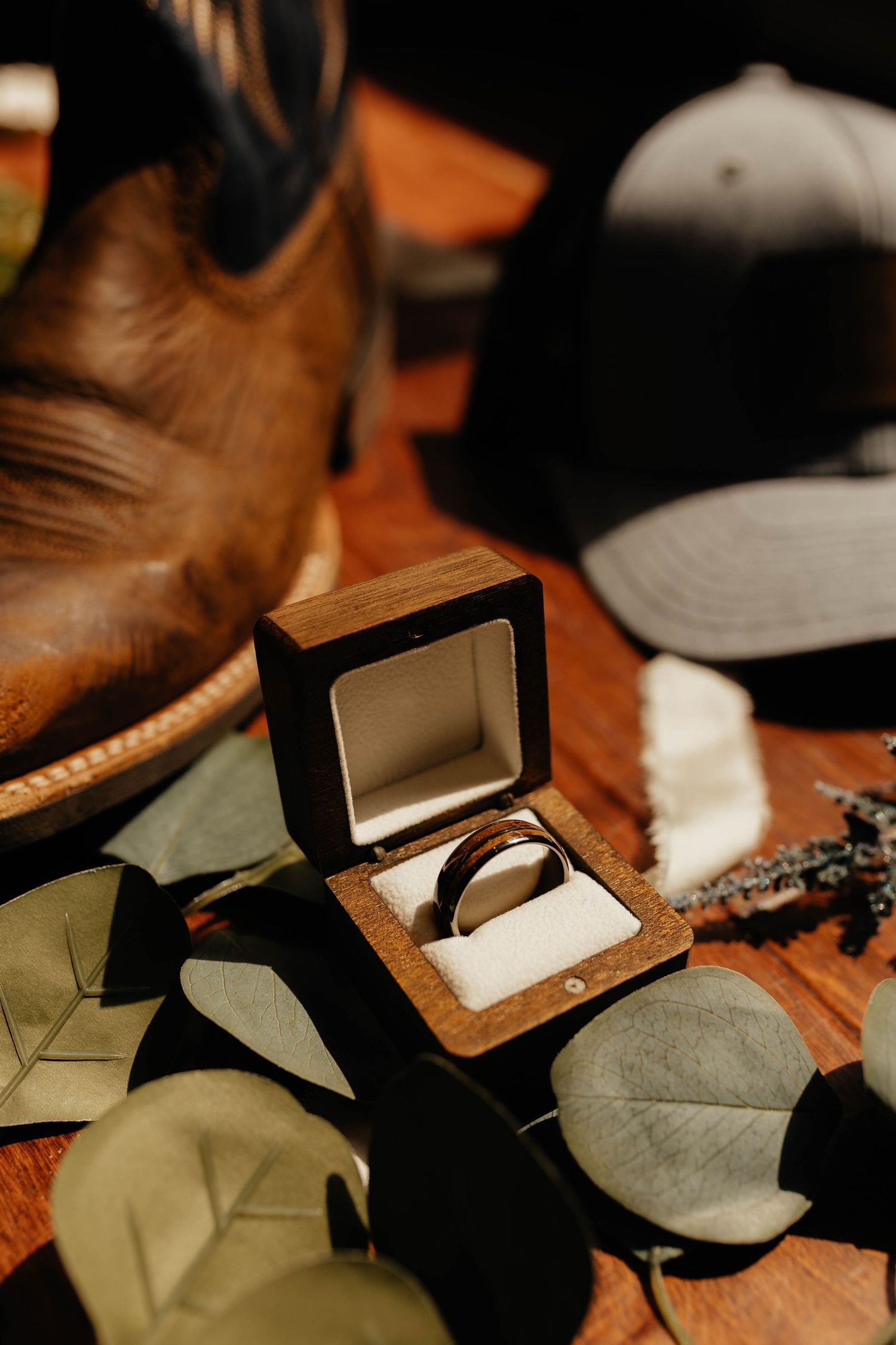 Wedding ring in wooden box surrounded by eucalyptus leaves, cowboy boot, and cap on rustic wood surface.