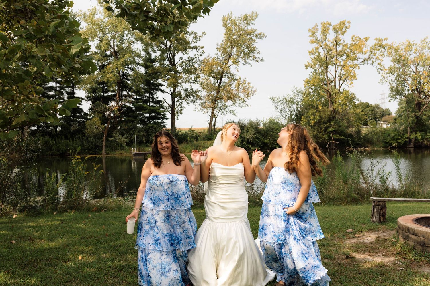 Bride in white gown walks joyfully with two bridesmaids in blue floral dresses by a lakeside.
