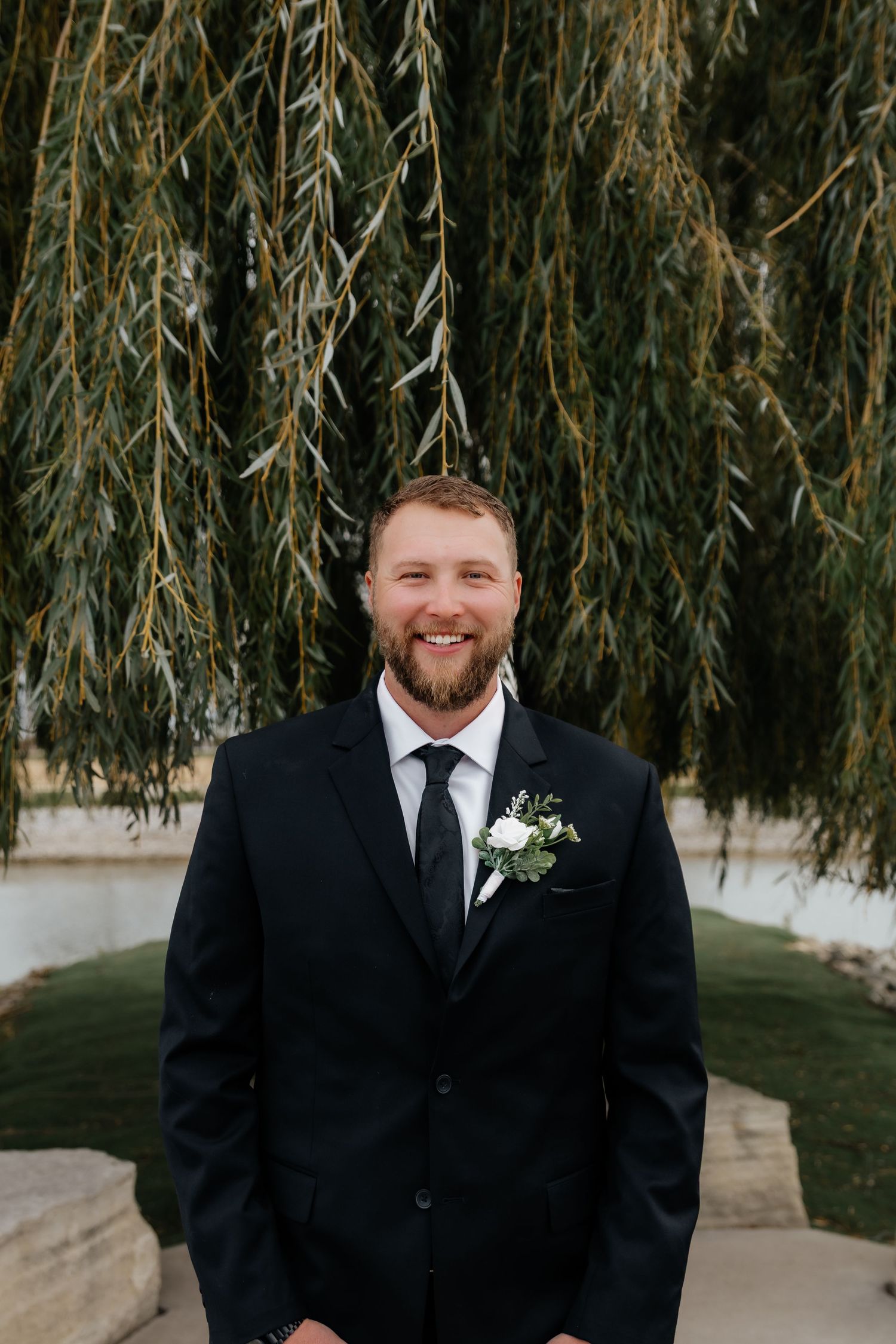Smiling groom in black suit with white boutonniere standing under weeping willow tree.