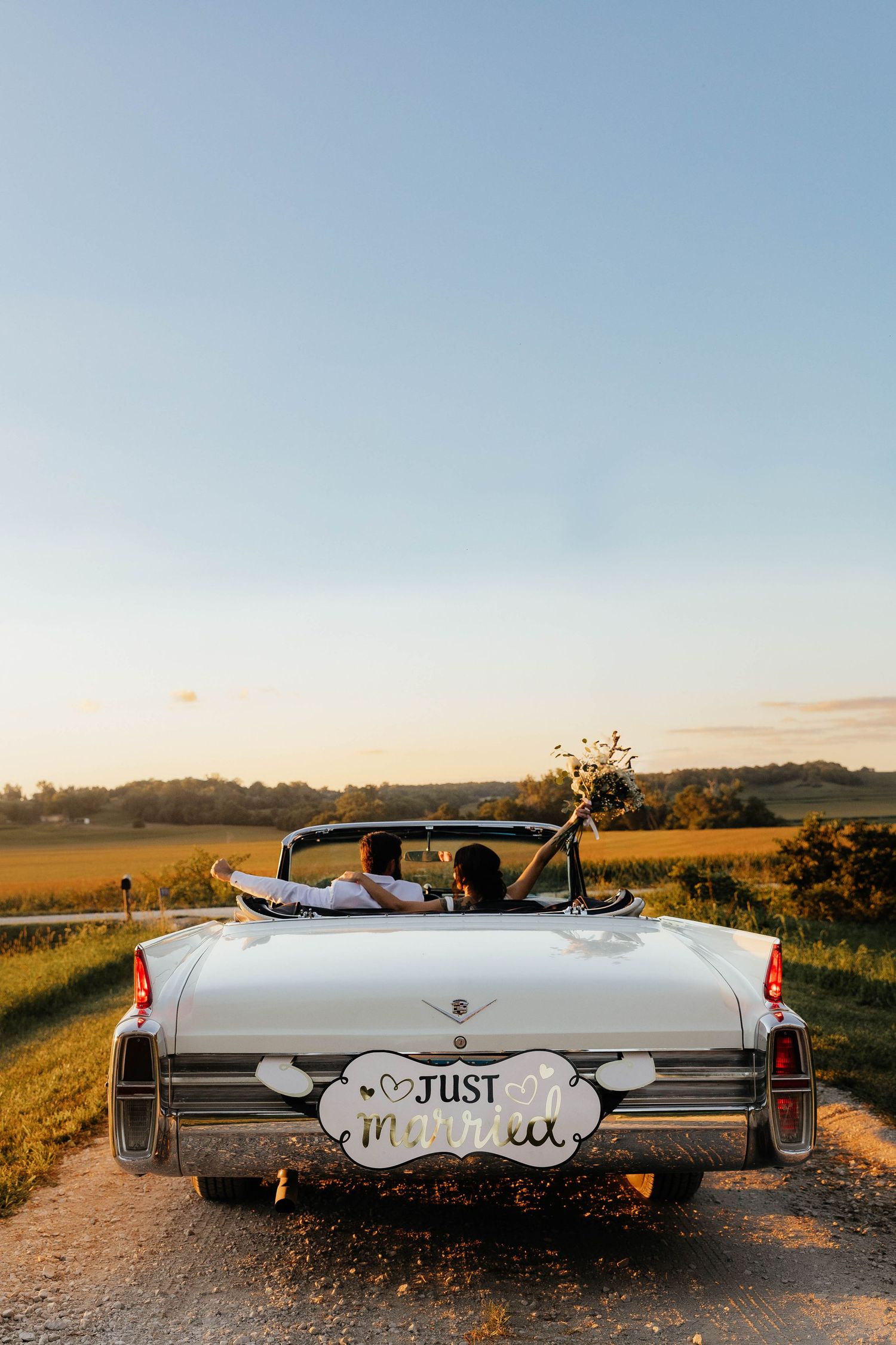 Newlywed couple driving away in a classic white convertible with Just Married sign on a scenic country road at sunset.