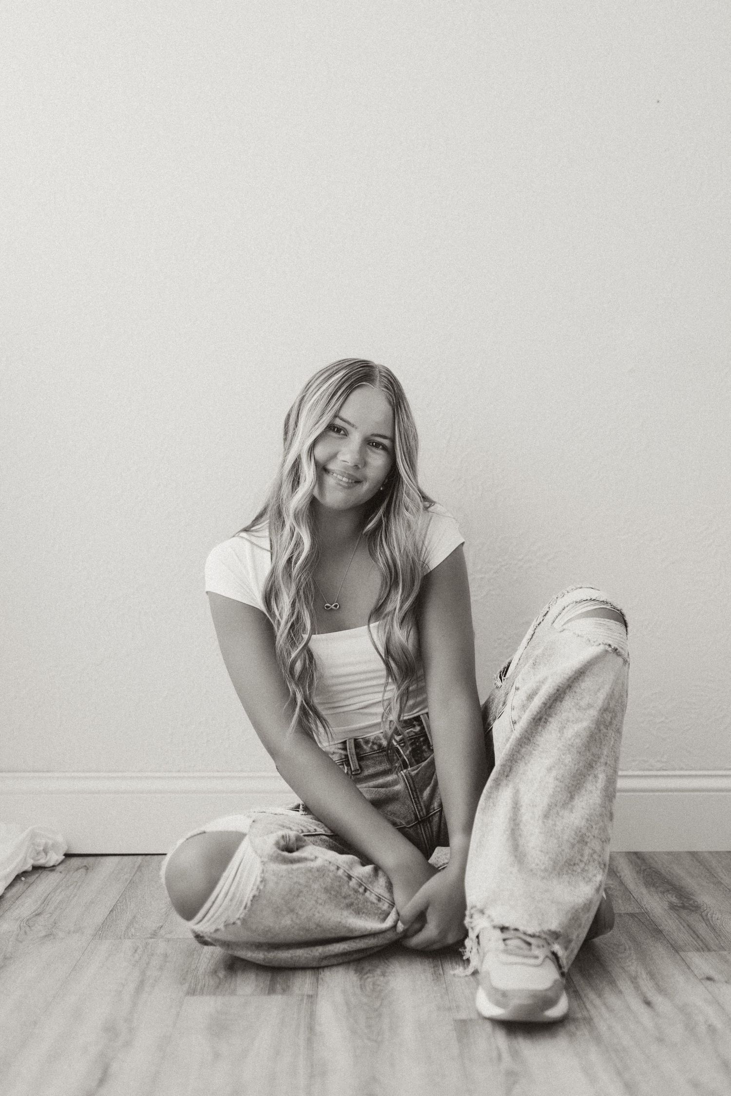 Black and white photo of a teenage girl with long blonde hair sitting on a wood floor in ripped jeans and white top.