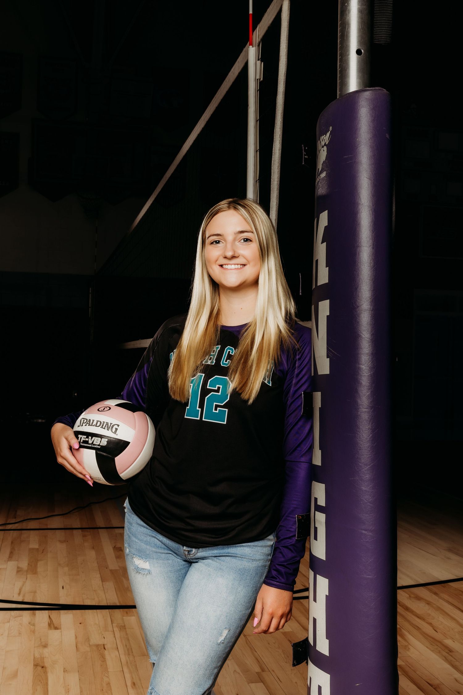 Blonde volleyball player in jersey number 12 leaning on net post holding a Spalding ball.