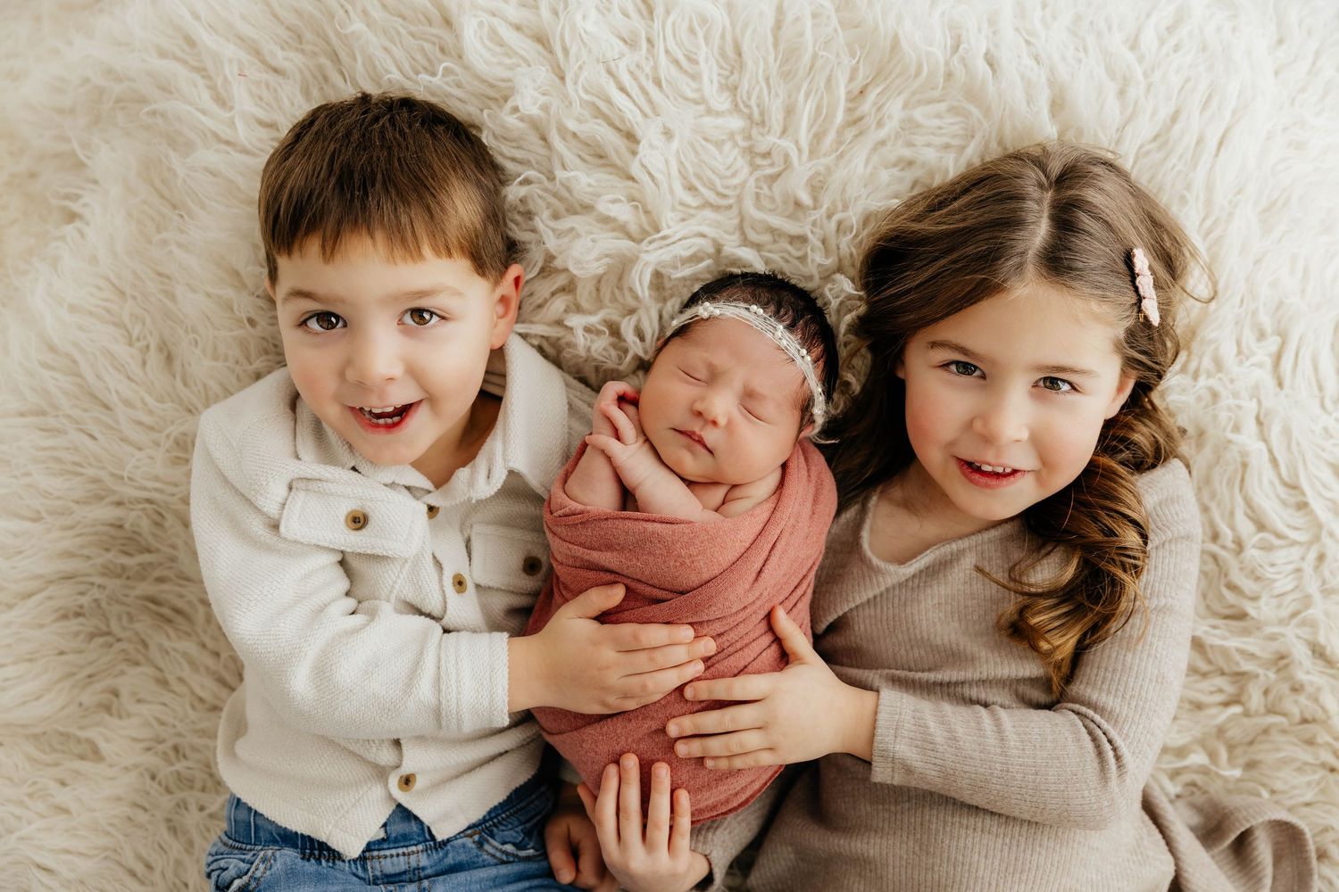 Two siblings smile holding their swaddled newborn baby sister on a fluffy cream rug.