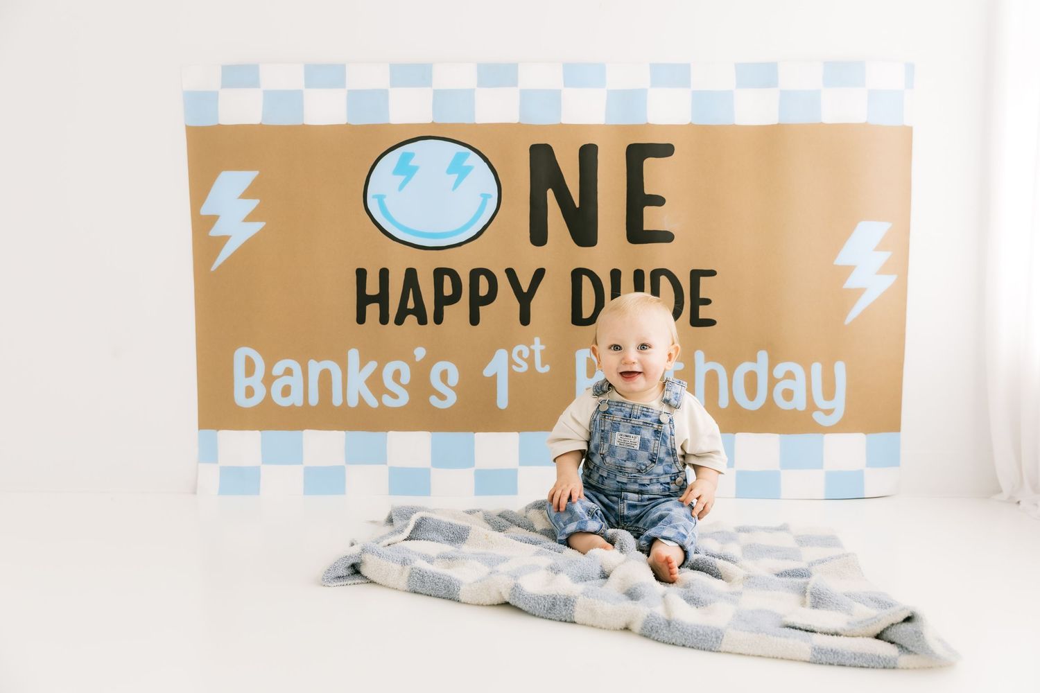 Smiling baby boy sitting on blanket in front of One Happy Dude first birthday sign.