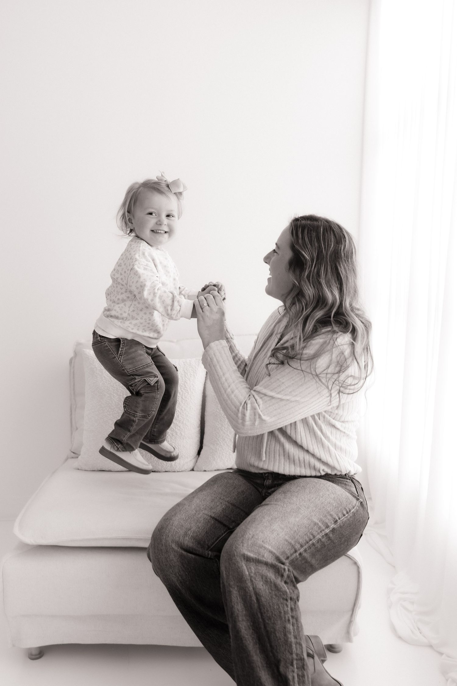 Black and white photo of mother holding hands with toddler standing on white studio bench.